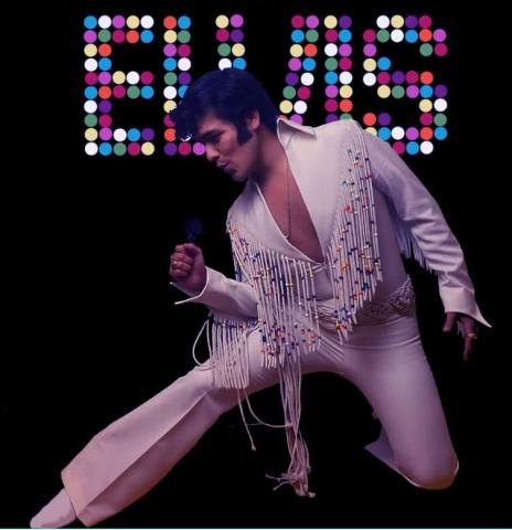 A man in a white Elvis-style jumpsuit with colorful fringe performs with a microphone.
