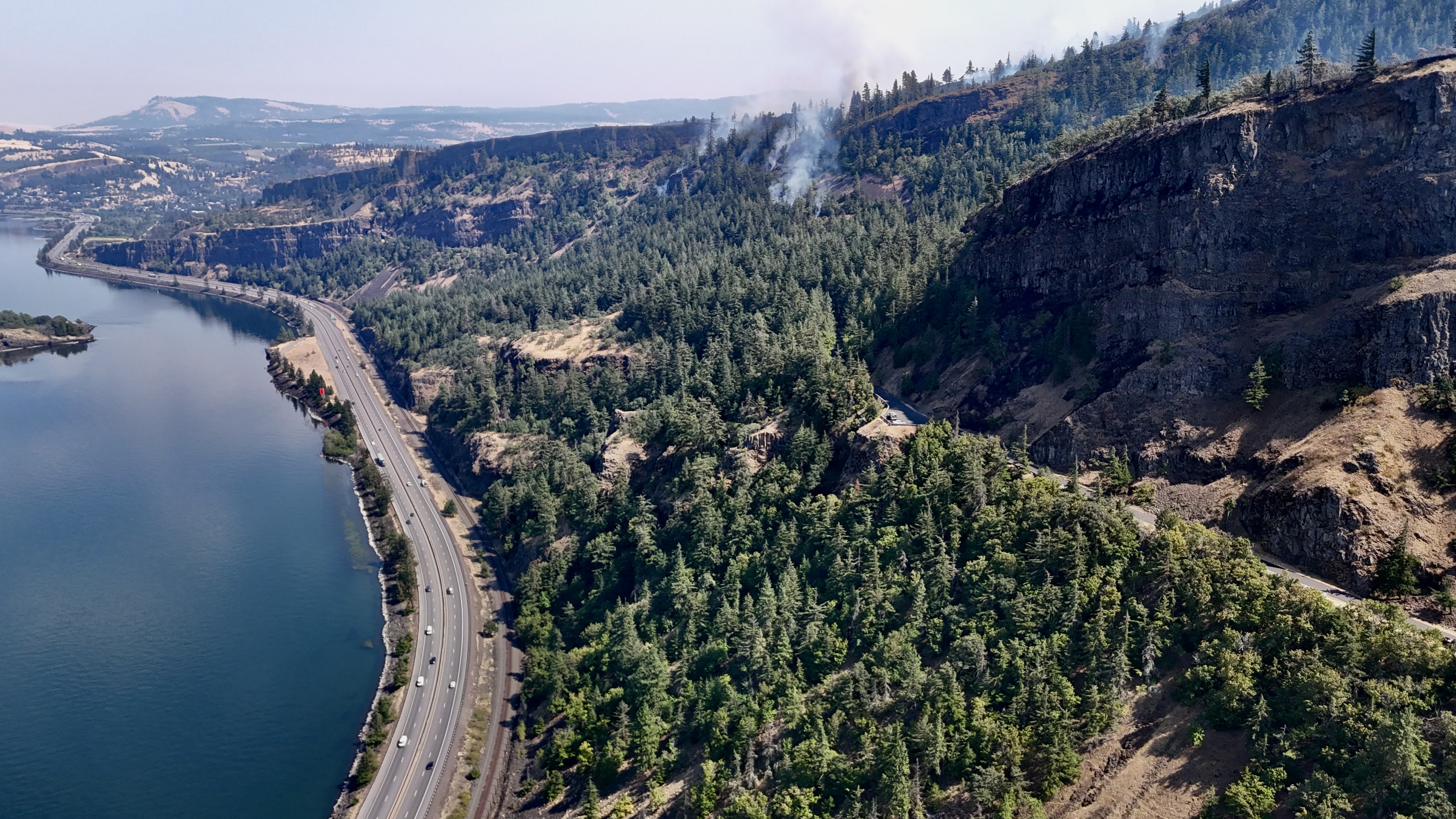 Aerial view of a forested river canyon with smoke rising from trees and a highway below.