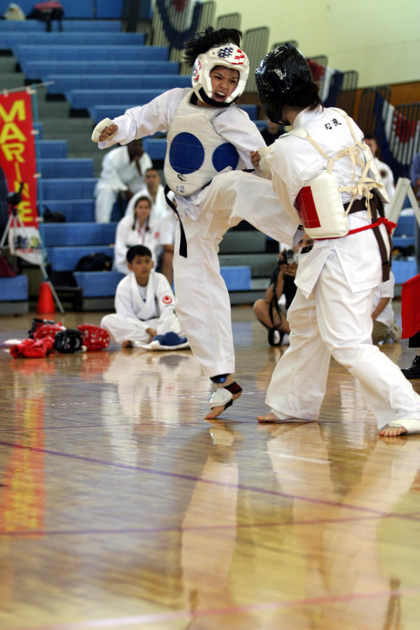 Two young karate competitors in white uniforms and headgear sparring at a tournament.