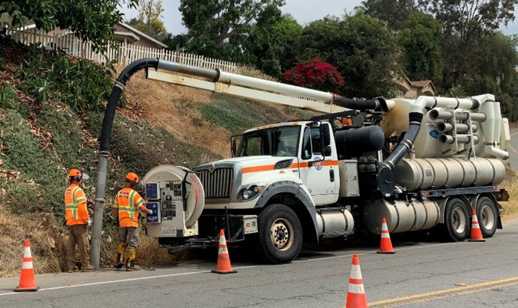 Workers in orange high-visibility vests operate a large utility truck on a suburban road.