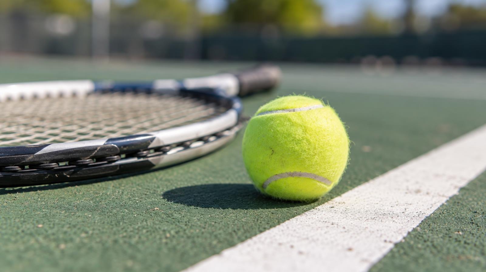 Yellow tennis ball and racket resting on a green hard court beside a white baseline.