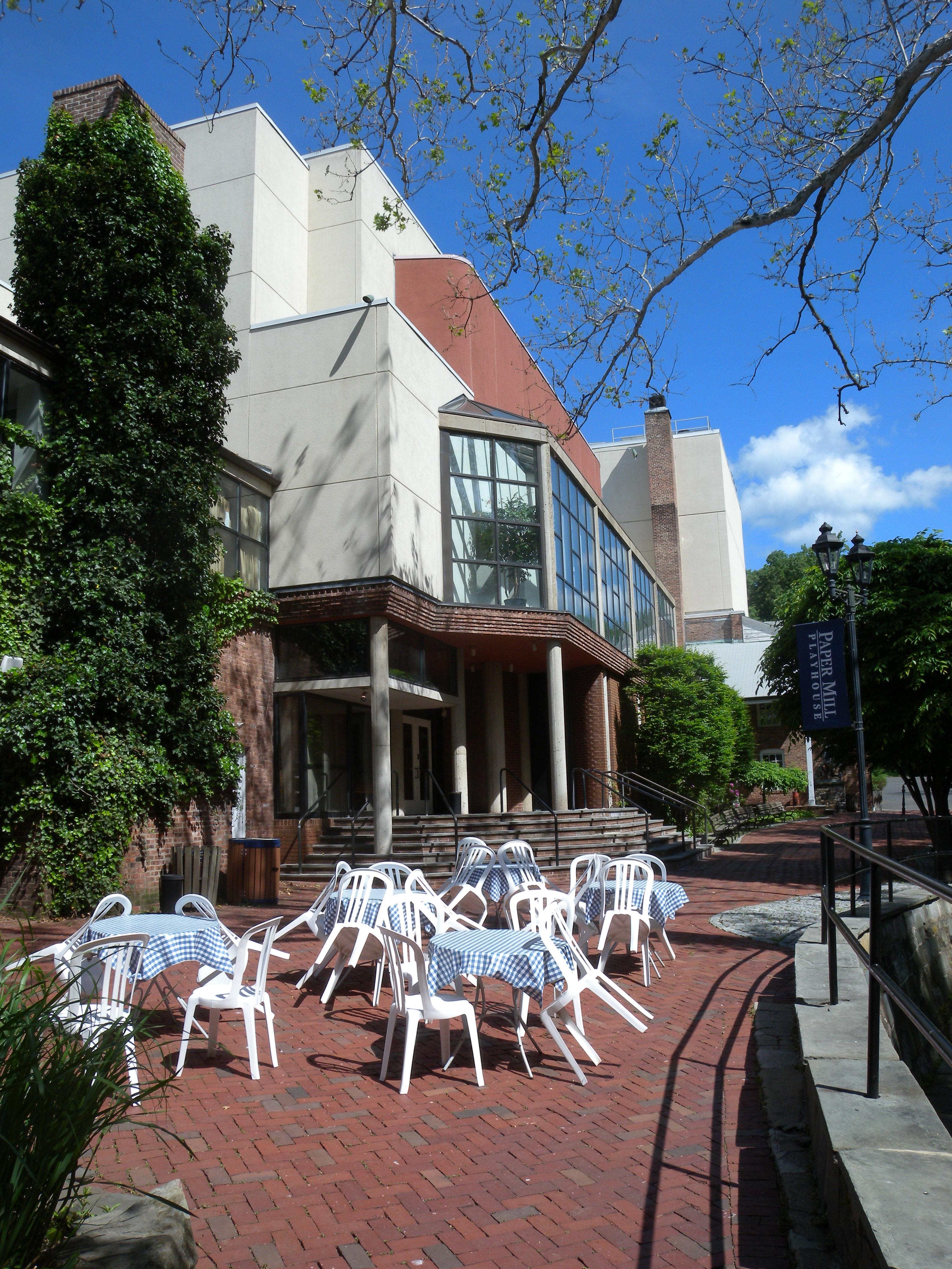 Outdoor terrace with white chairs and checkered tablecloths in front of a theater building.
