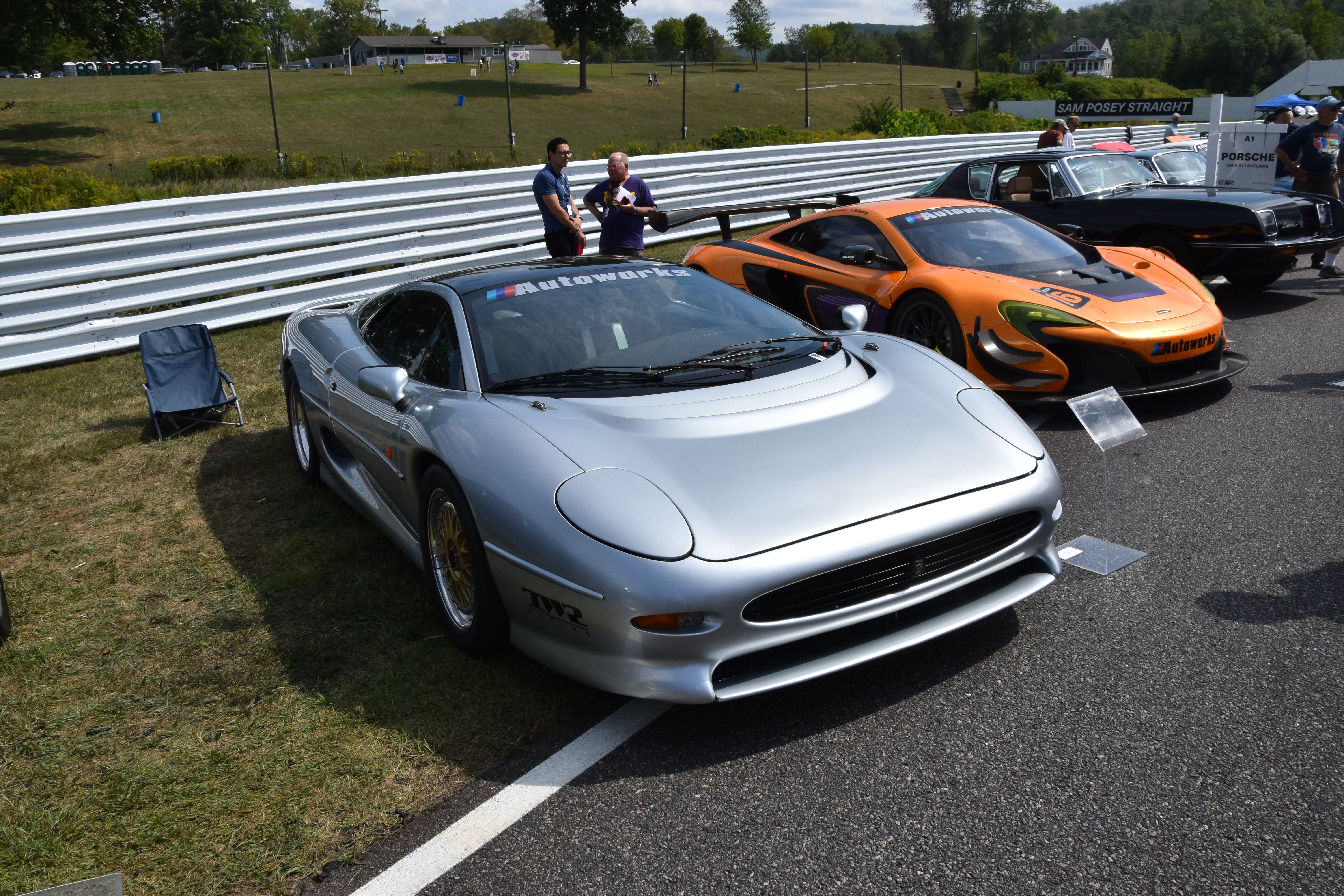 A silver sports car in the paddock at Lime Rock Park with track barriers and spectators visible.