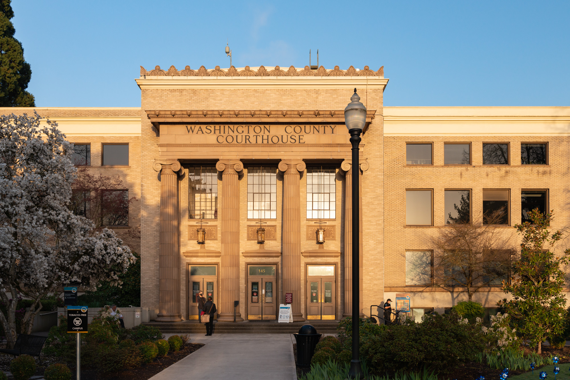 Neoclassical courthouse with columns and inscribed name, flanked by blooming trees at golden hour.