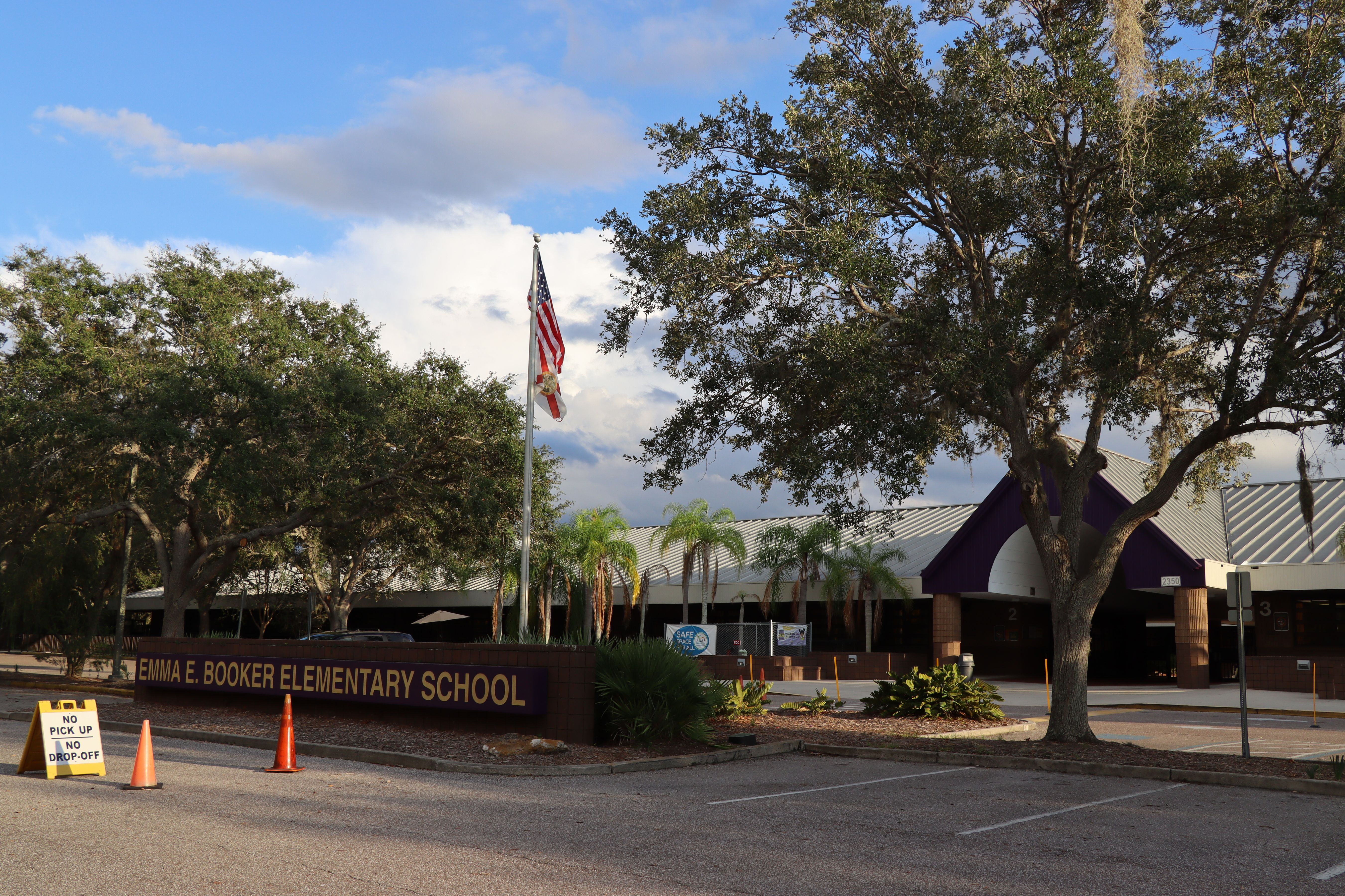 Front entrance of a school building with a sign, flagpole, and palm trees.
