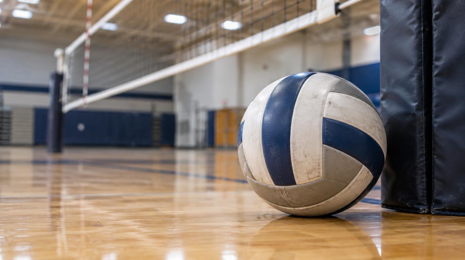 A volleyball resting on a hardwood gymnasium floor near the net post.