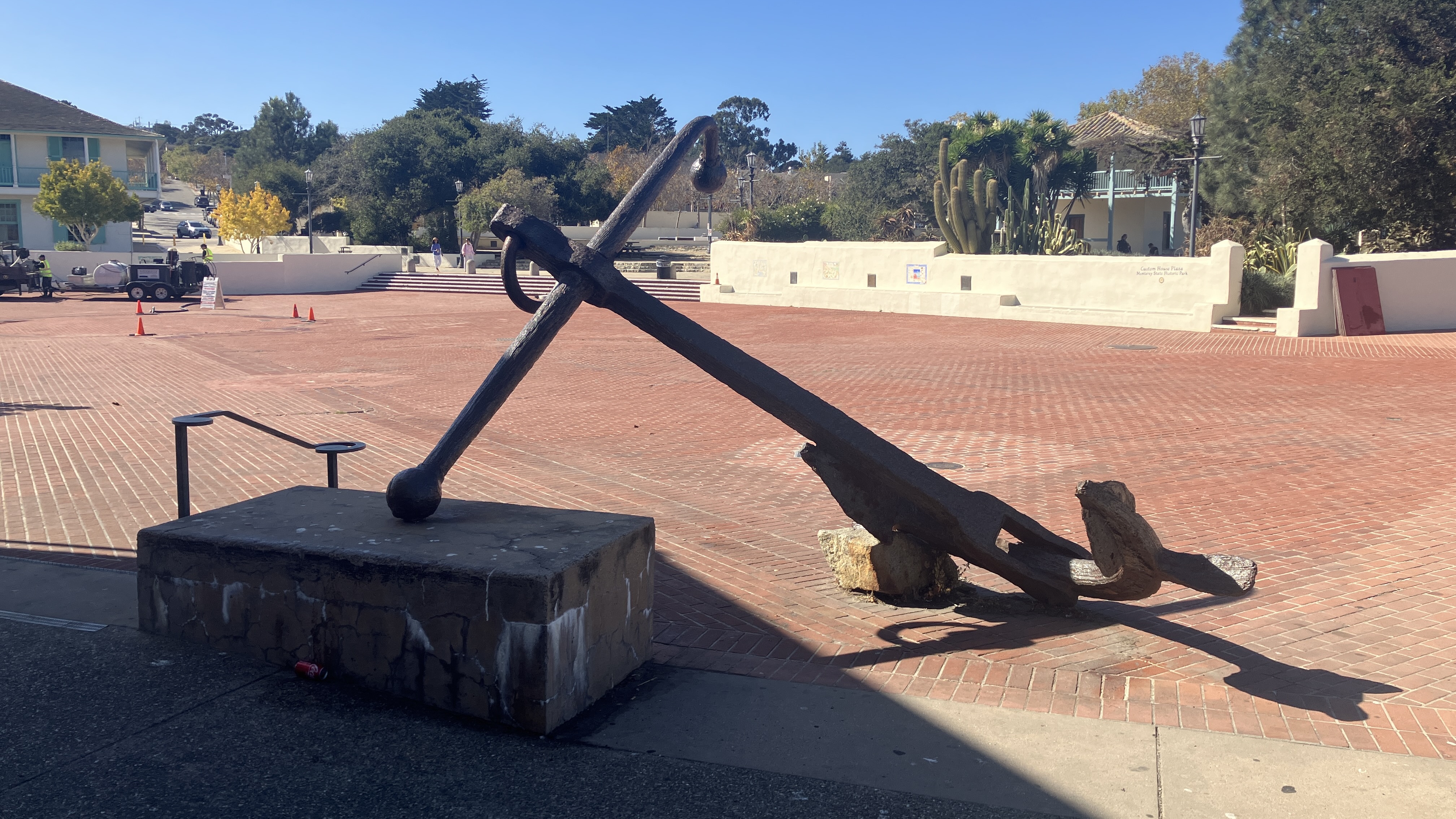 Large iron ship anchor on a stone pedestal in an open brick plaza on a sunny day.