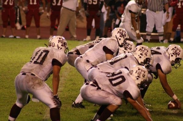 High school football players in white uniforms line up at the line of scrimmage under stadium lights at night.