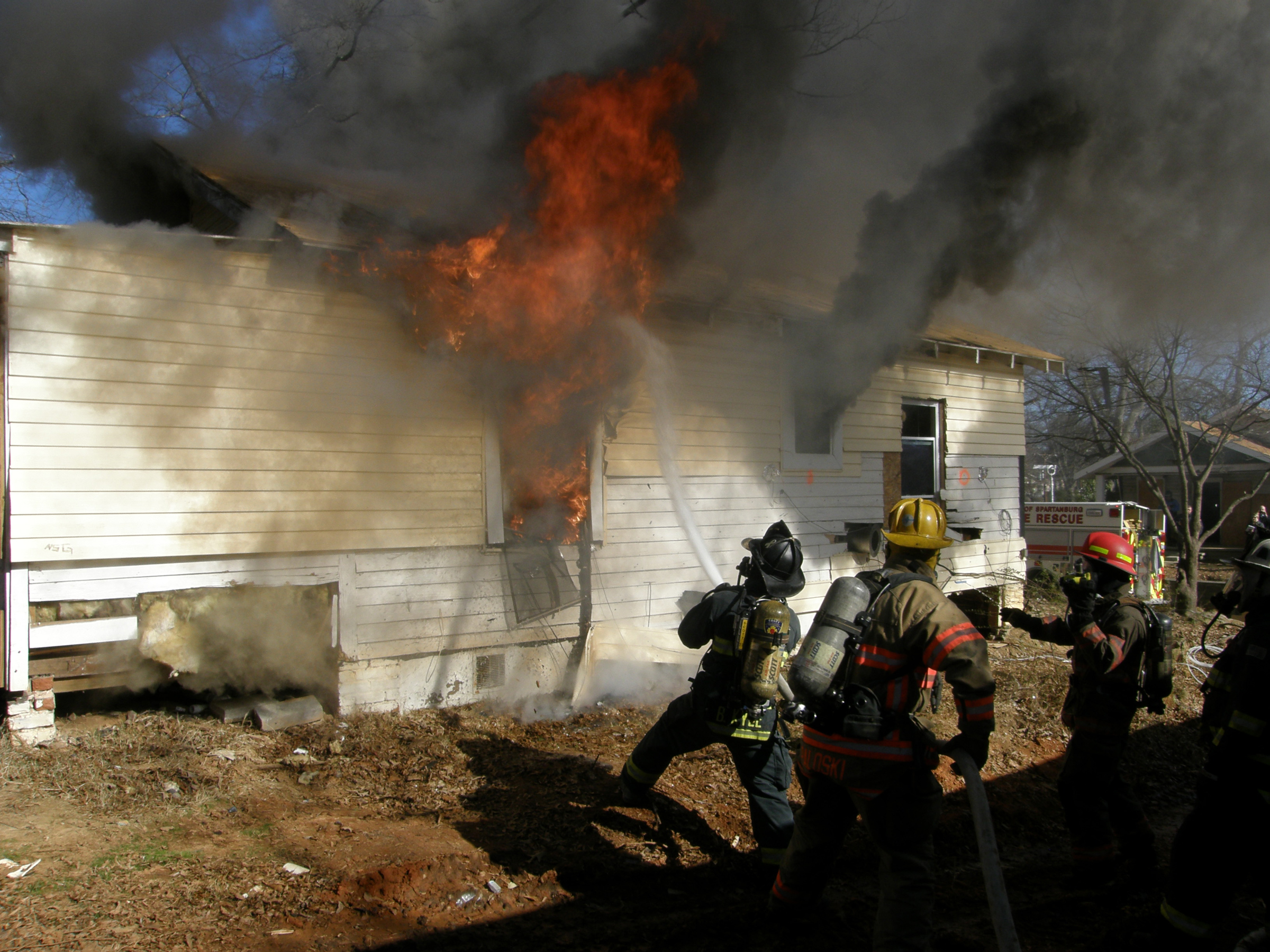 Firefighters in full gear approaching a burning single-story house with heavy flames and black smoke.