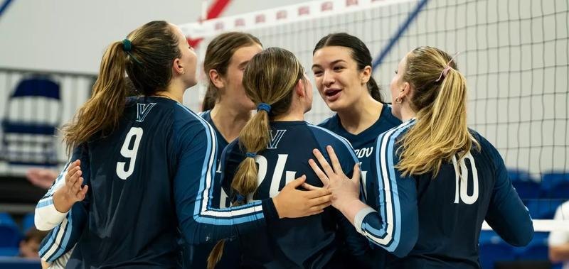 Five volleyball players in navy uniforms huddle together on an indoor court.