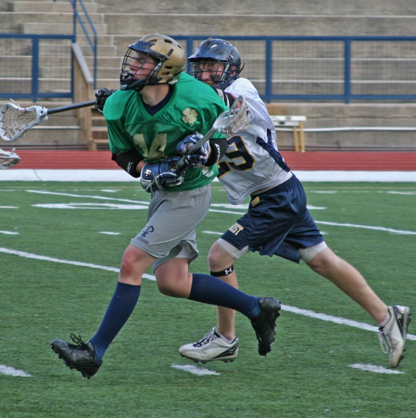 Two high school lacrosse players in action on a turf field, one carrying the ball while being pursued by a defender.