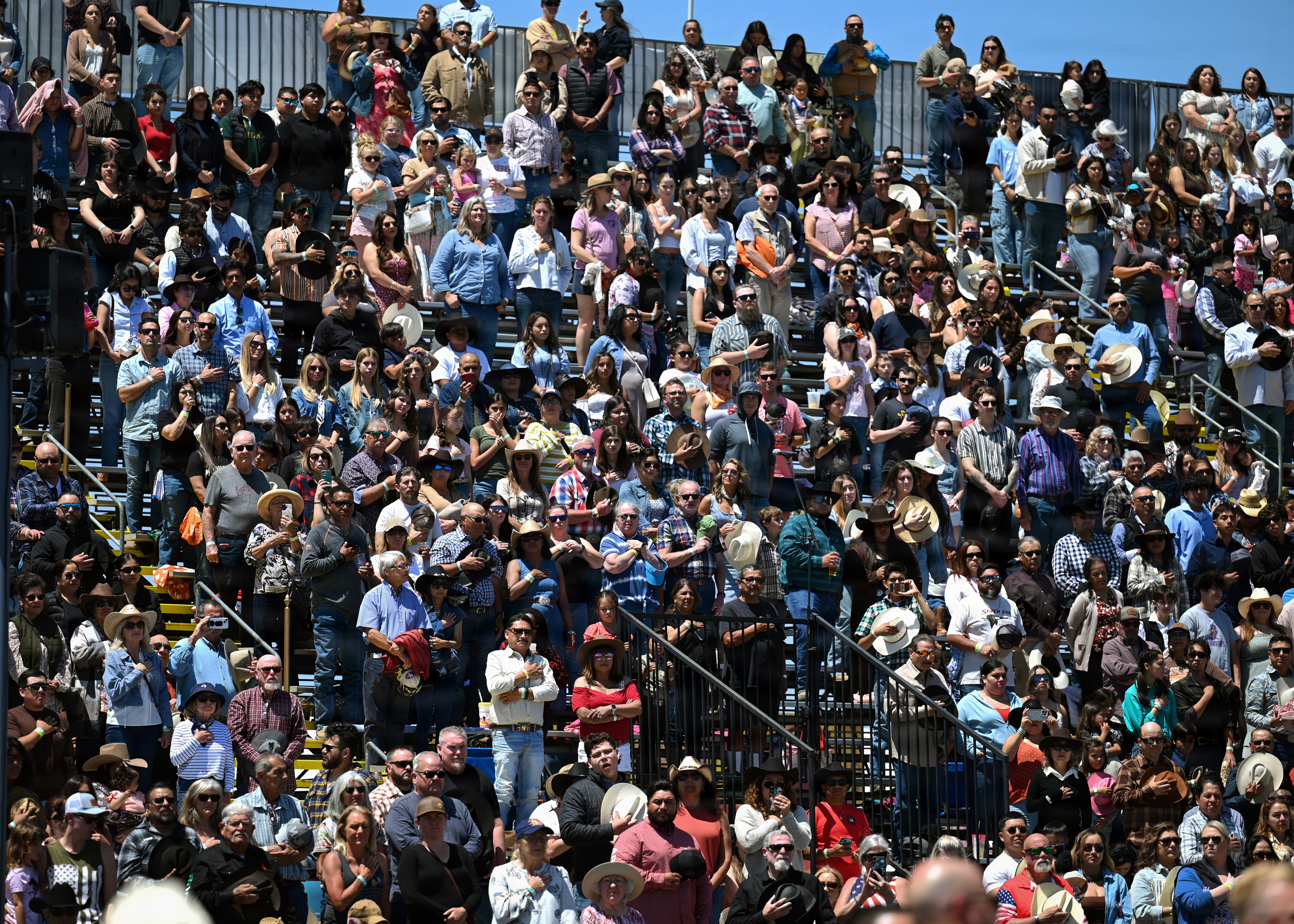A large crowd in cowboy hats fills bleacher seating at an outdoor rodeo arena on a sunny day.