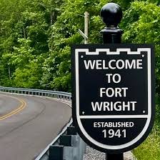 Black shield-shaped welcome sign reading 'Welcome to Fort Wright – Established 1941' beside a tree-lined road.