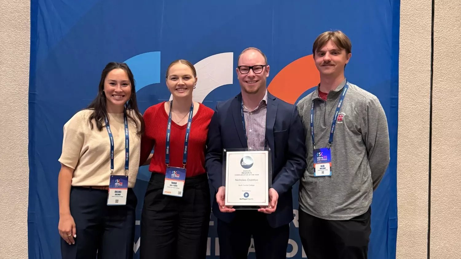 Four people wearing conference lanyards pose together, one holding a framed award, in front of a blue backdrop.