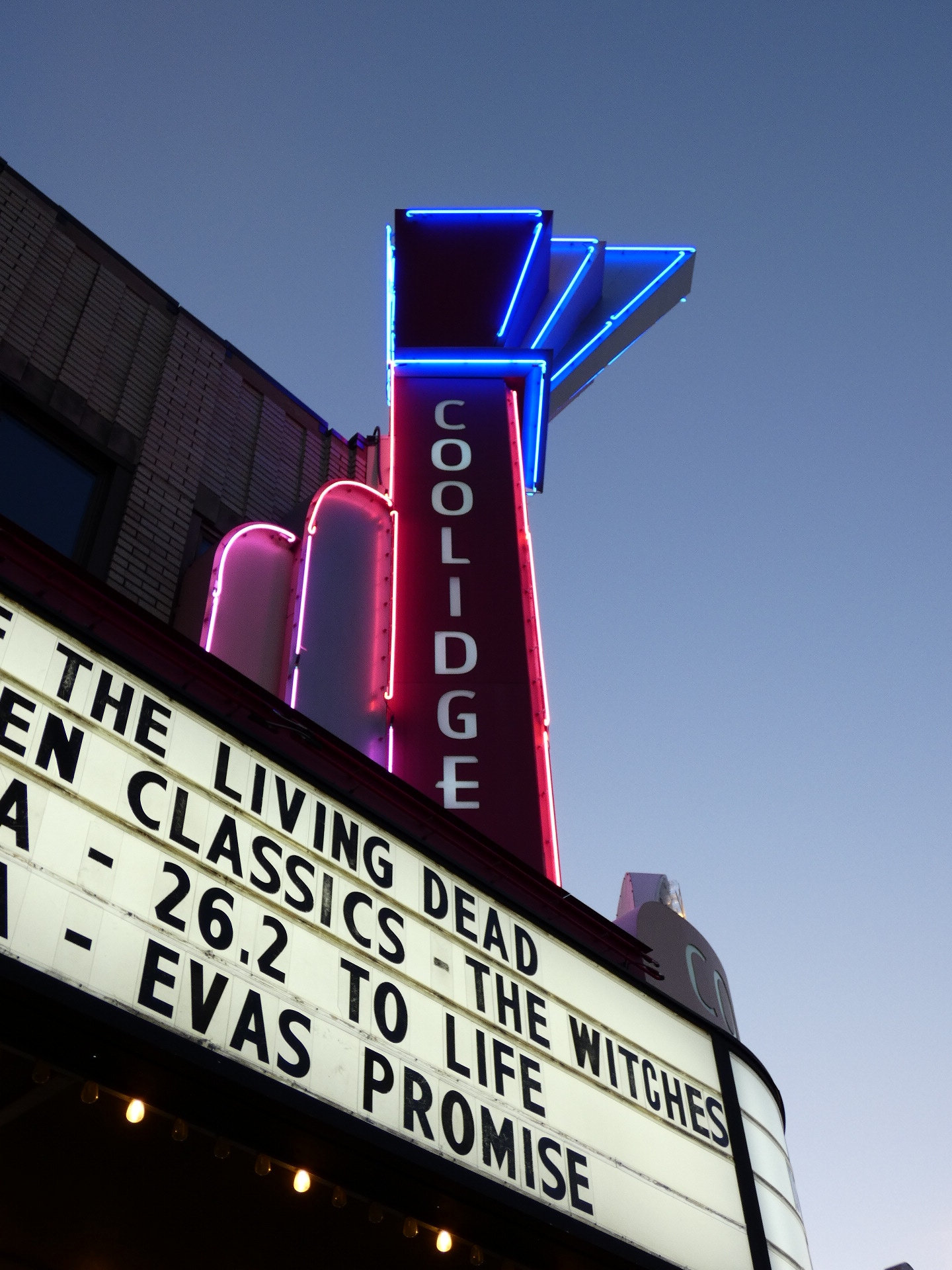 Neon-lit theater marquee and vertical sign glowing at dusk against a blue-grey sky.