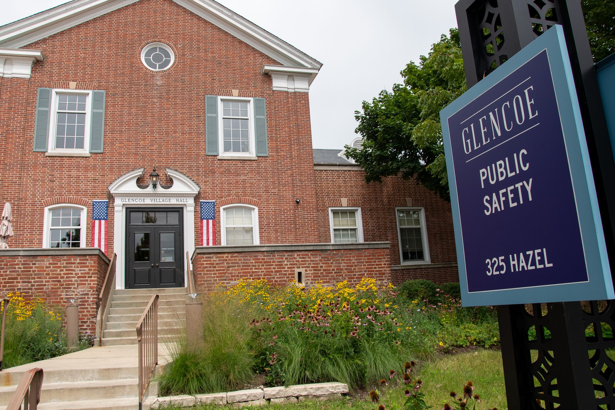 Red brick village hall building with American flags and a public safety sign in front.