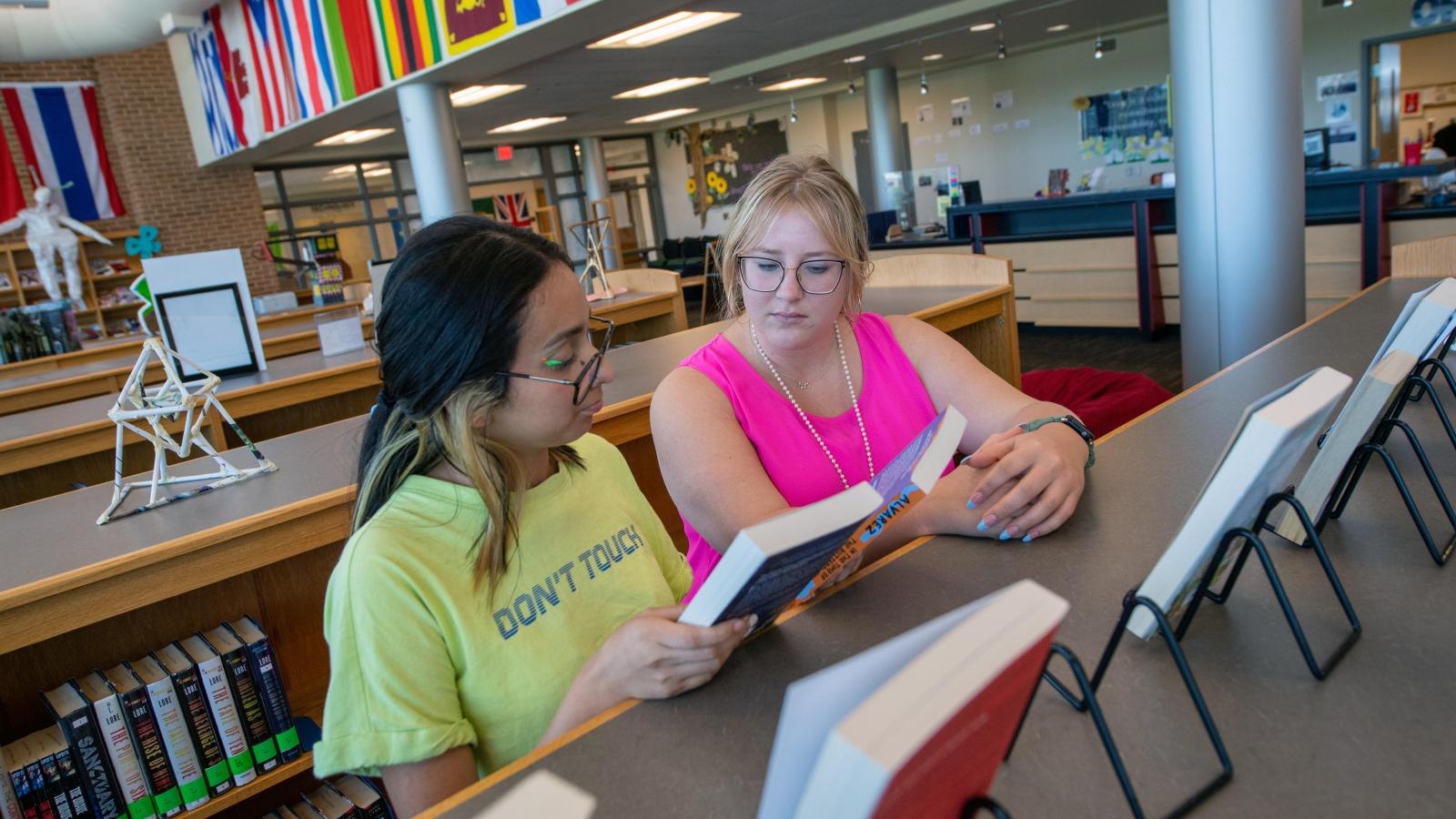 Two students wearing glasses read and discuss a book at a table in a school library with international flags overhead.