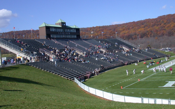 College stadium with bleacher seating, a press box, and colorful autumn foliage behind.