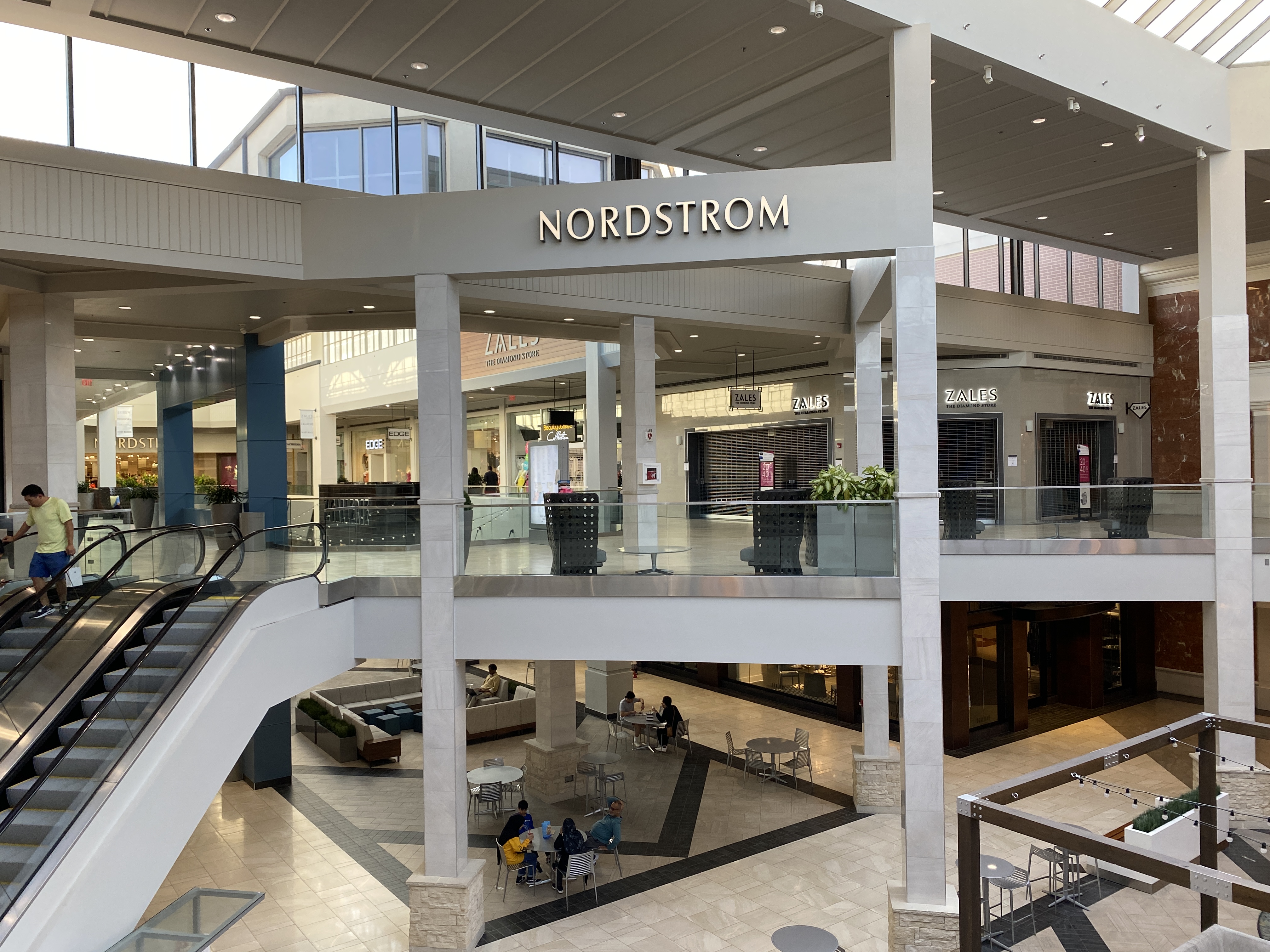 Multi-level mall interior with escalators, glass railings, and a large anchor store entrance.