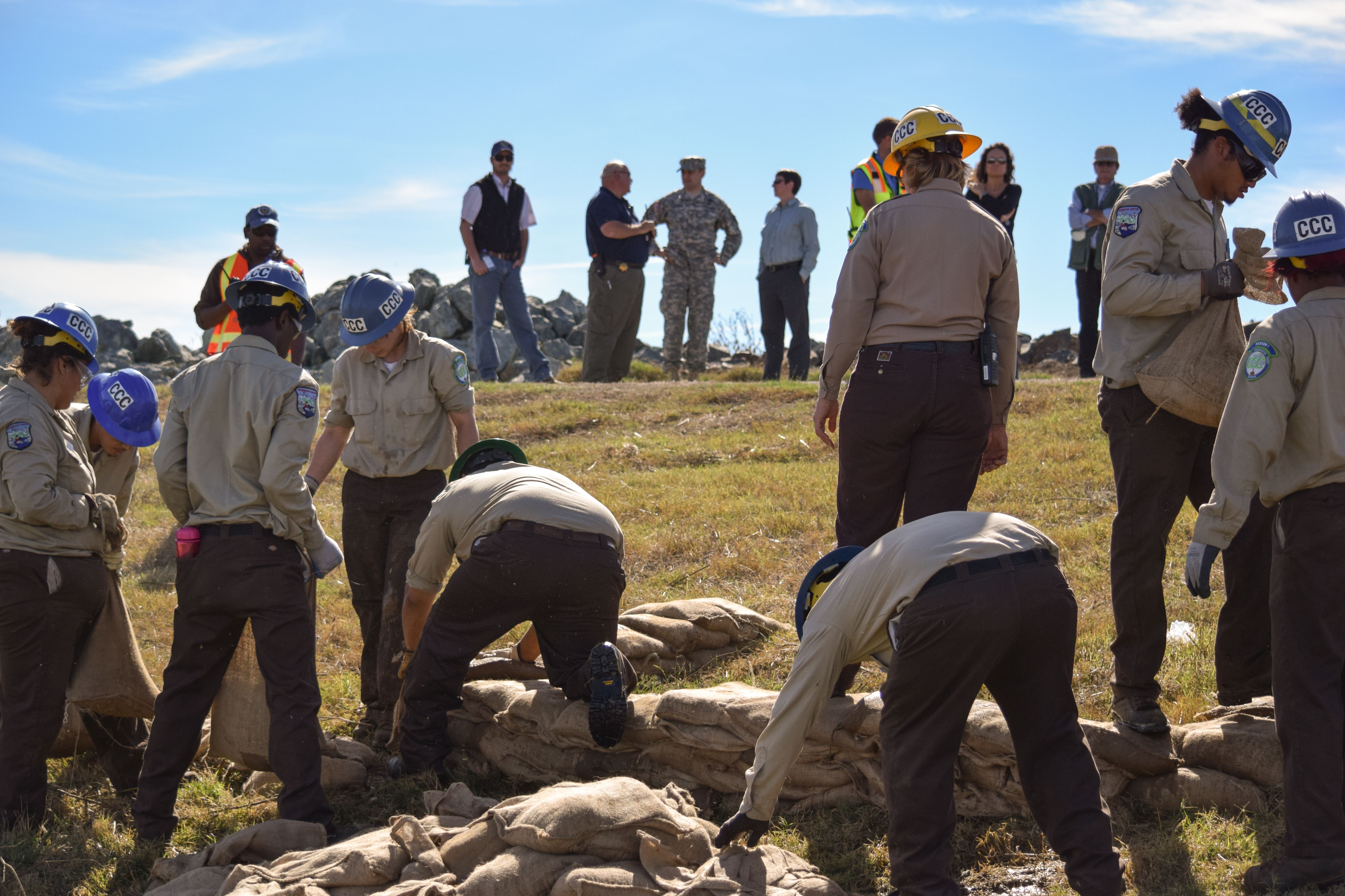Workers in blue helmets fill and stack burlap sandbags on a grassy field.