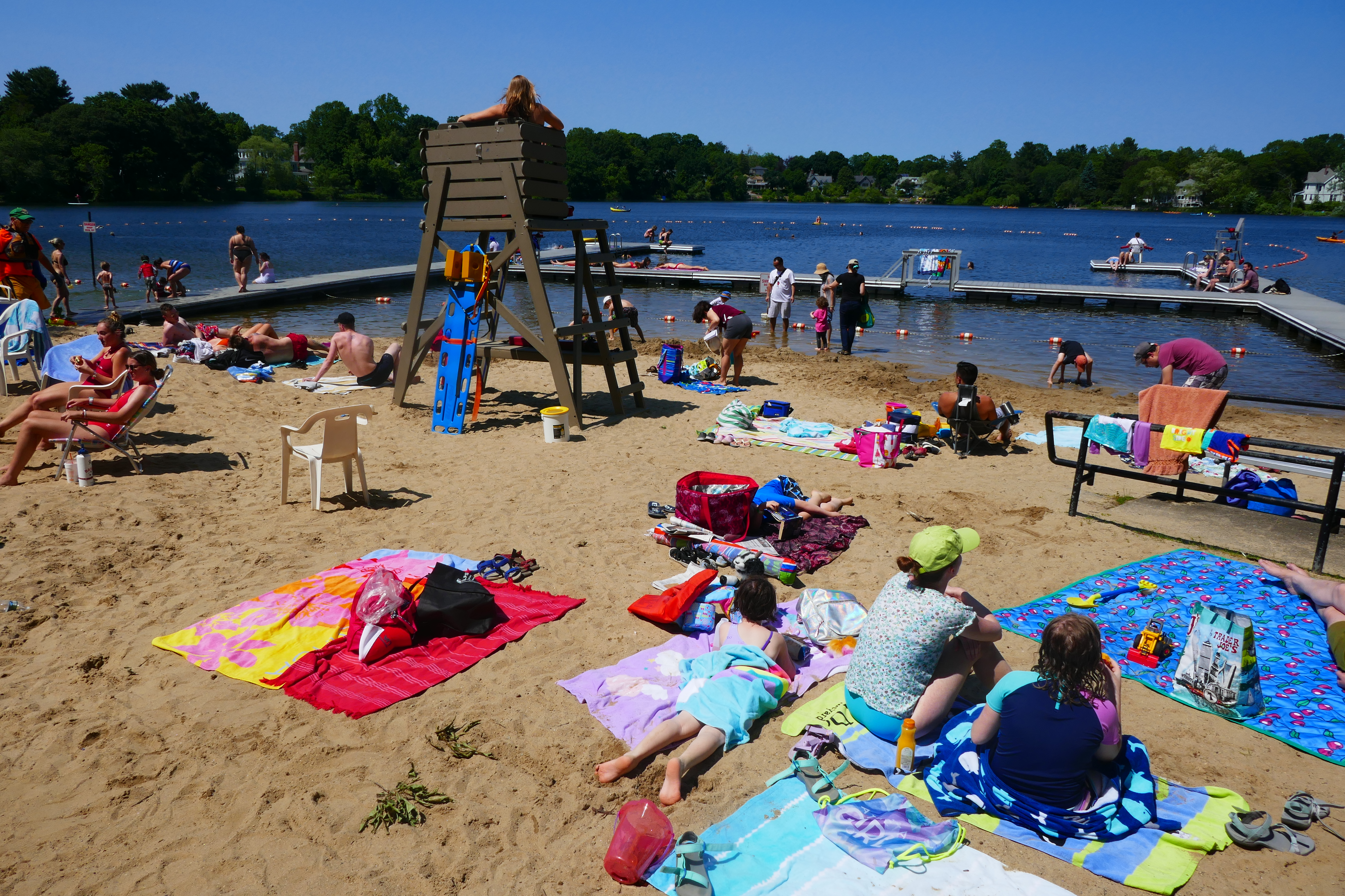 Crowded sandy beach at a lake with swimmers, a lifeguard stand, and a floating dock.