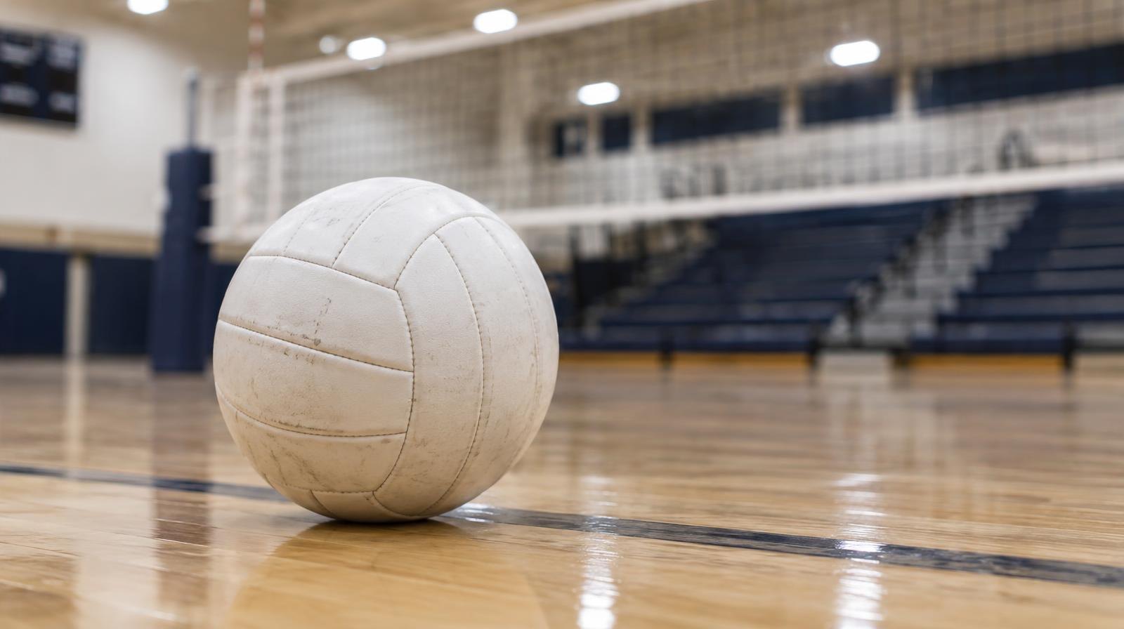 A white volleyball on a hardwood gym floor near a net with bleachers in the background.