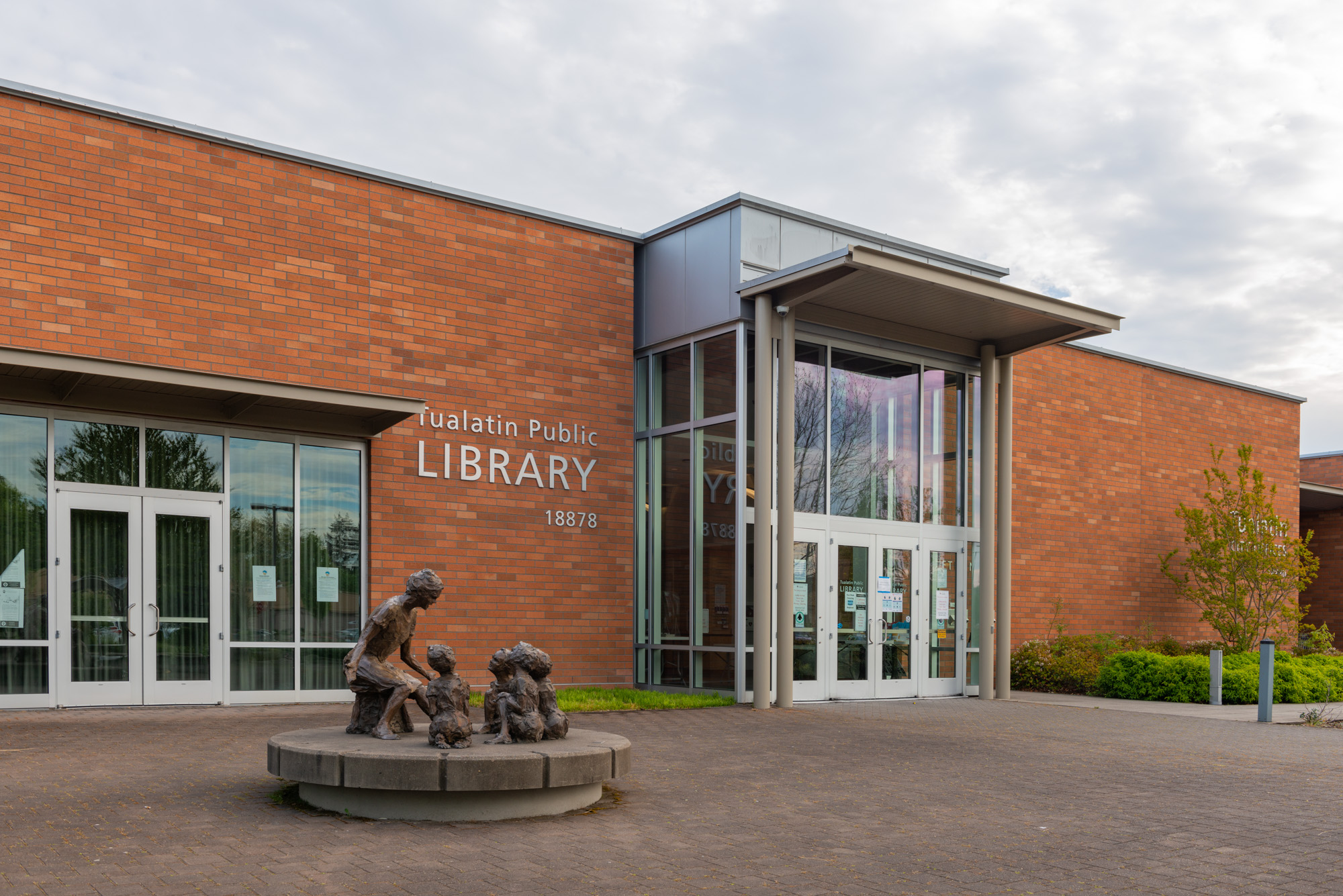 Red brick library entrance with glass doors and a bronze sculpture of a woman reading to children.