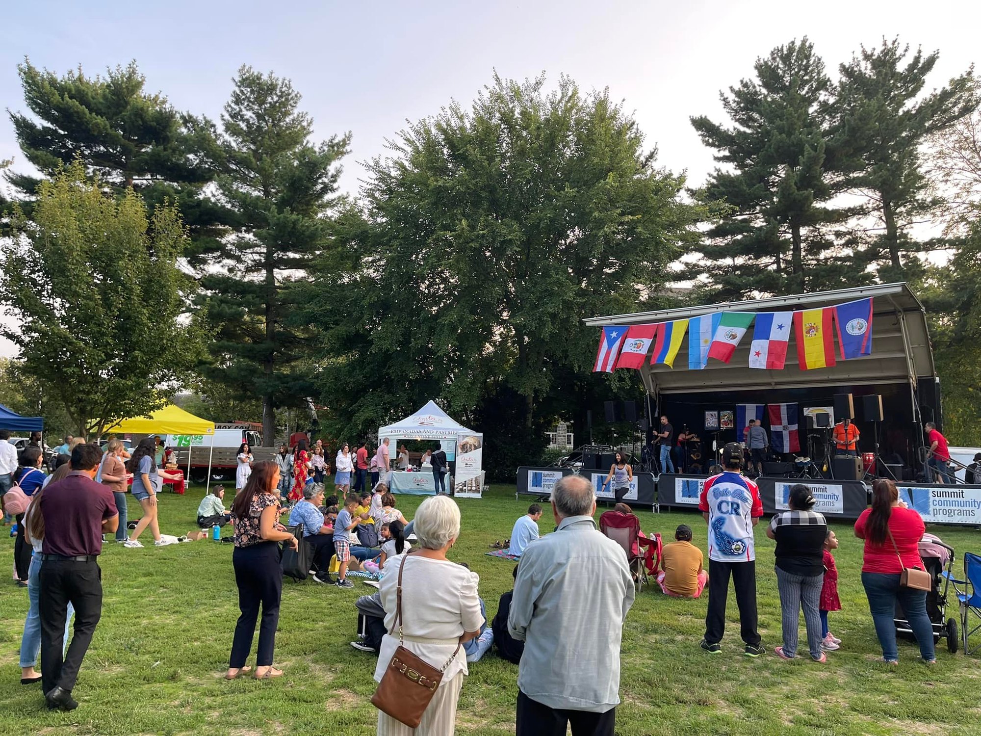 Crowd on a lawn facing an outdoor stage decorated with Latin American flags and vendor tents.