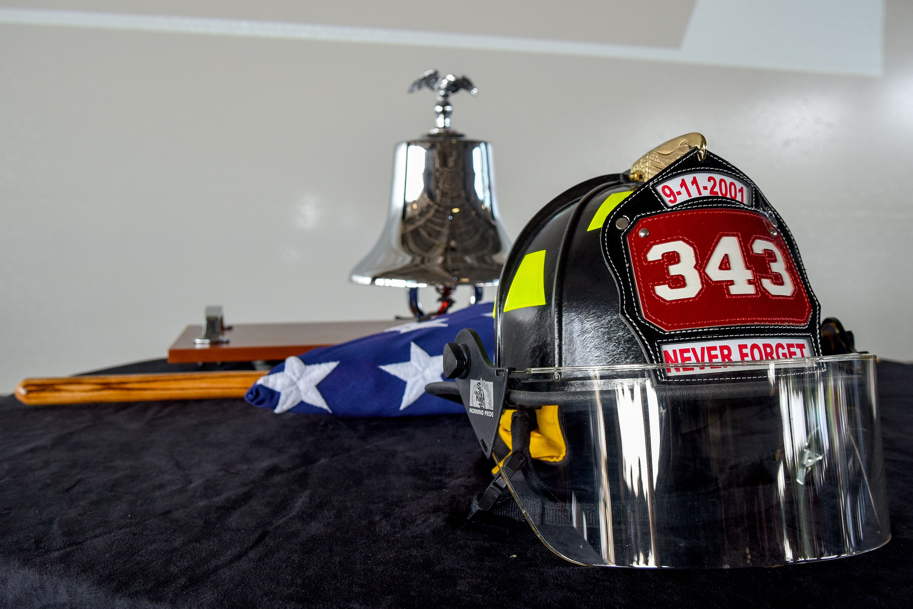 Firefighter helmet reading '343 Never Forget' beside a chrome bell and folded American flag.