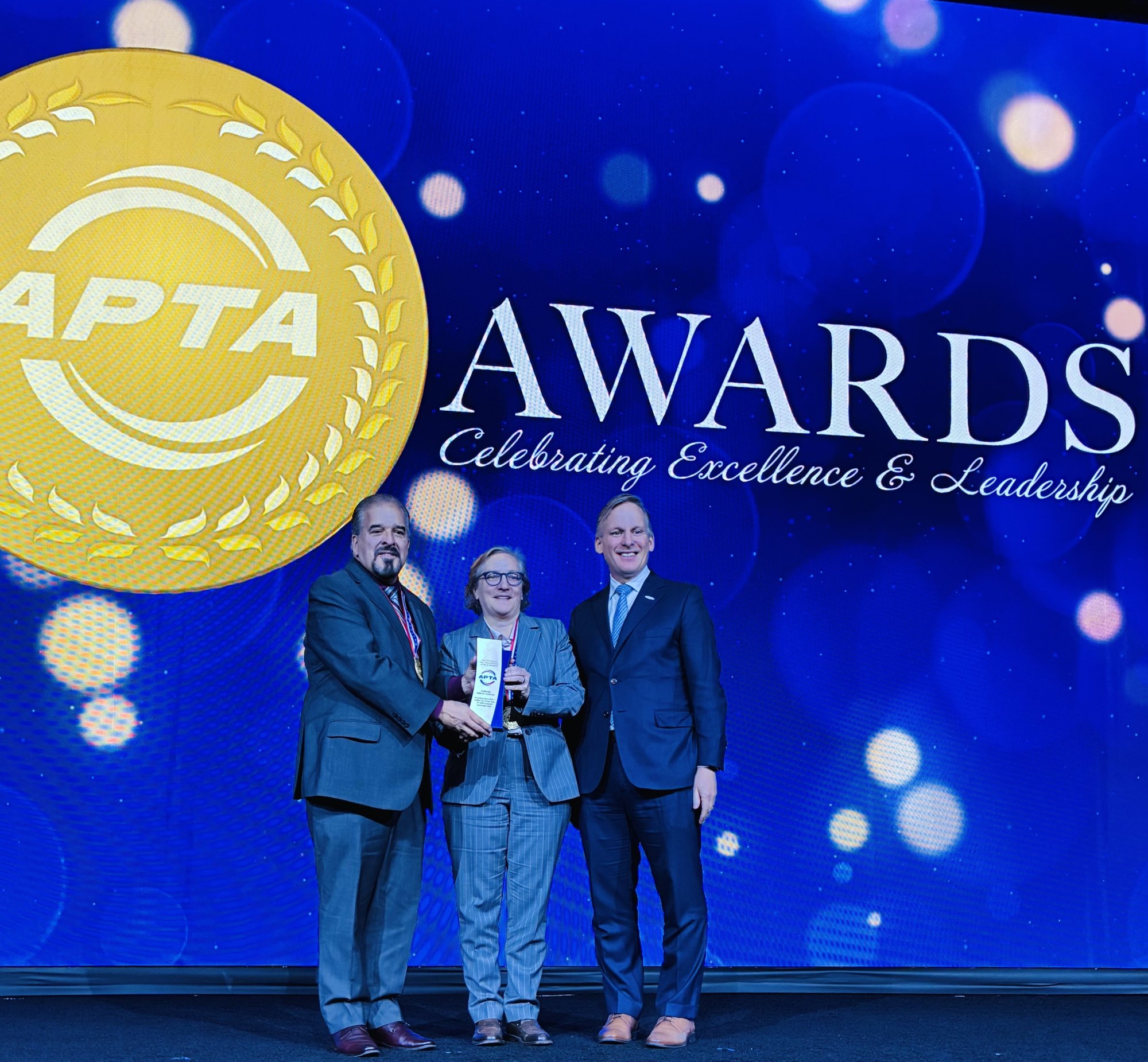 Three people in business attire stand on a stage holding a transit award in front of an APTA backdrop.
