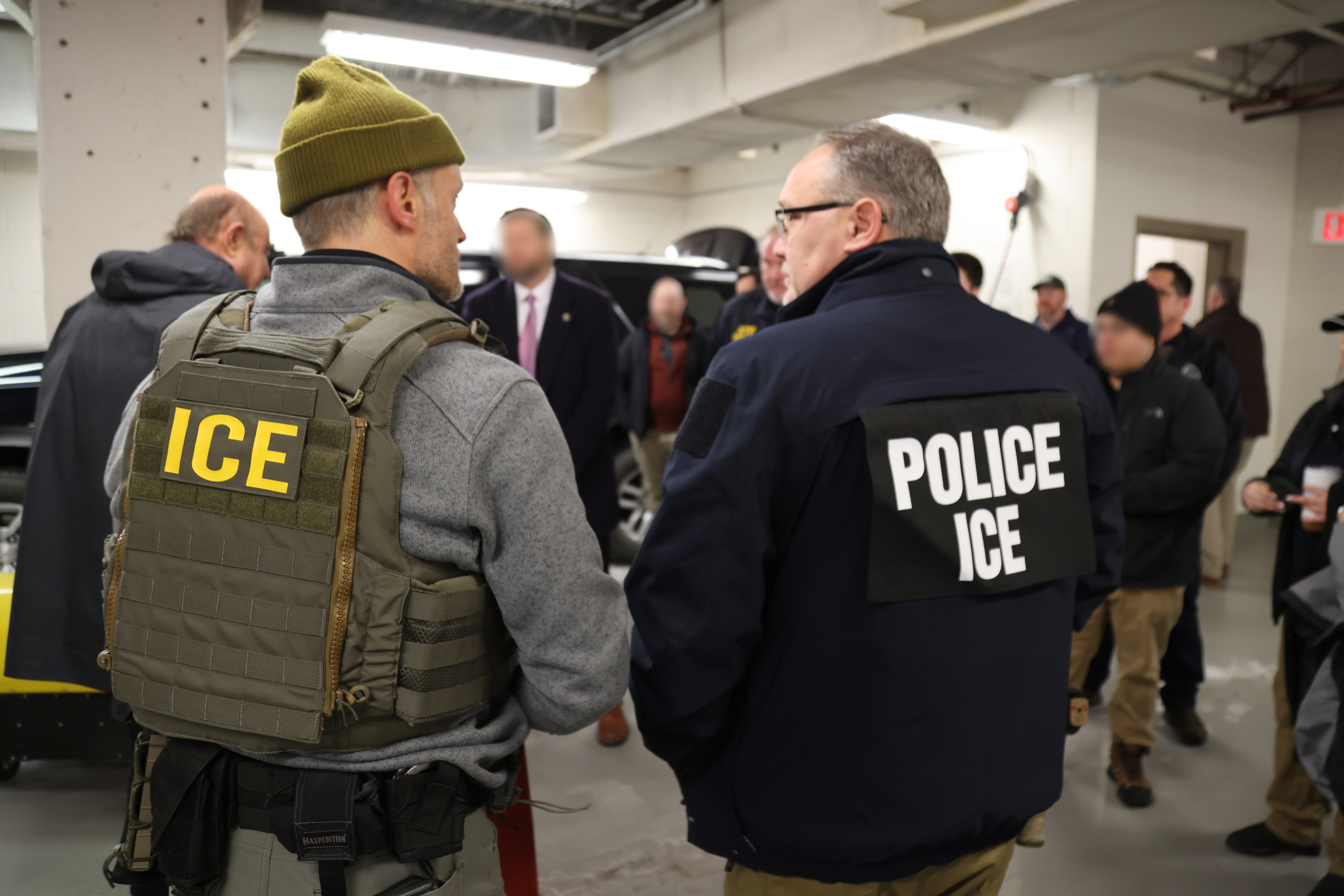 Two officers wearing ICE-labeled vests stand in a parking structure.