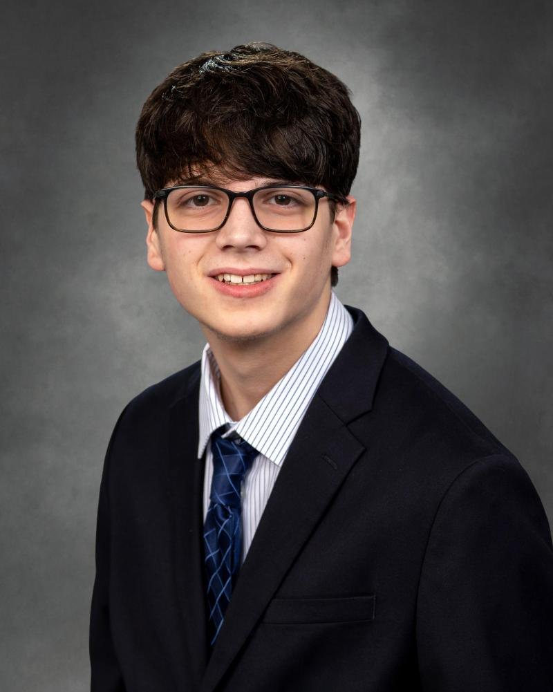 A young man in glasses, navy blazer, and striped tie poses for a formal portrait.
