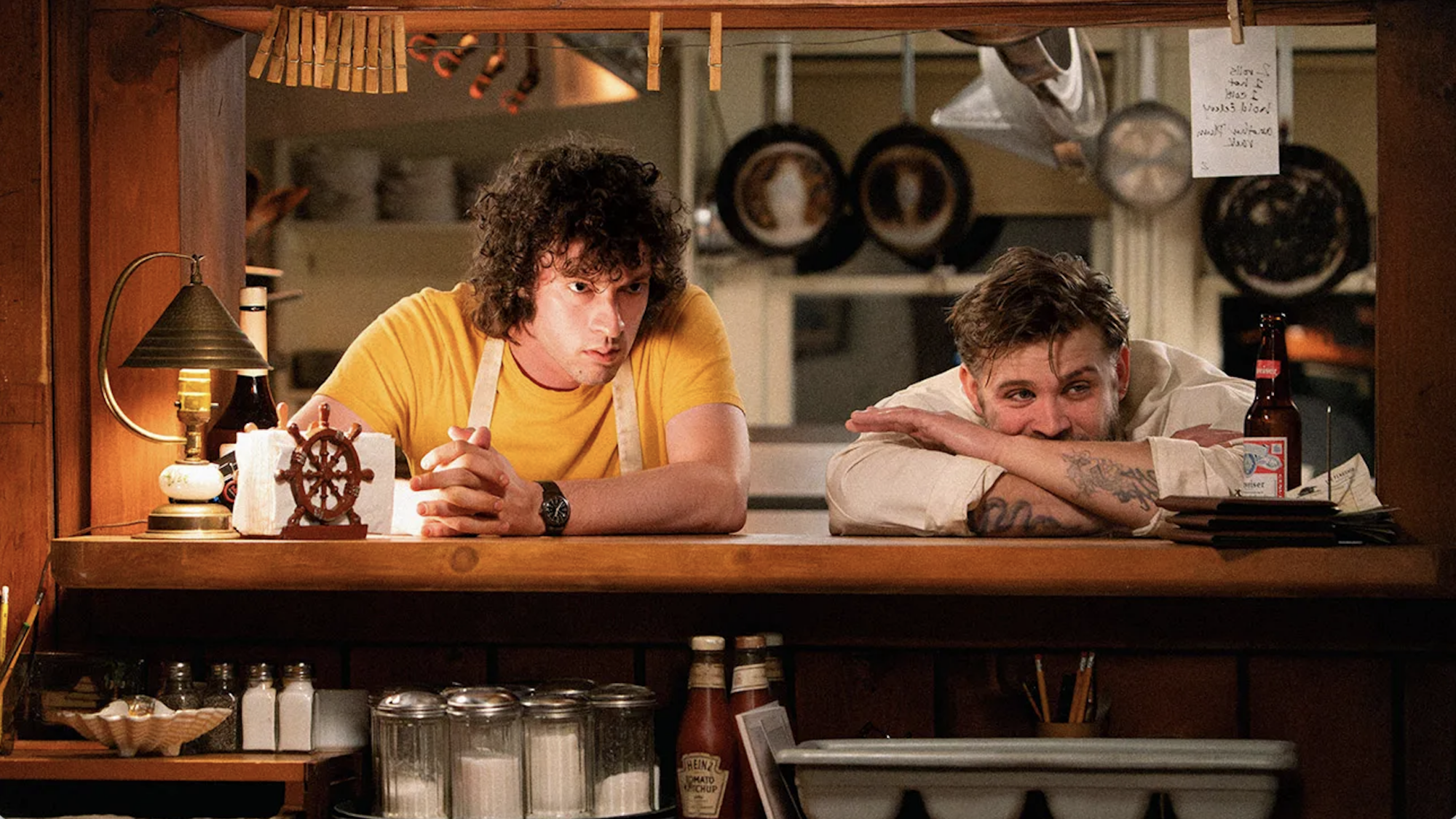 Two young men lean on a wooden diner counter surrounded by hanging pots and pans.
