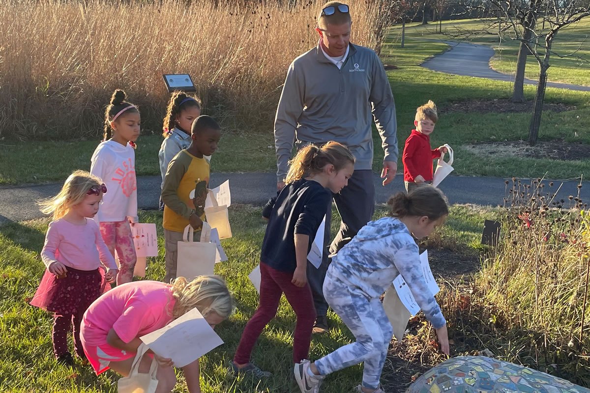 An adult leads a line of young children across a grassy park area outdoors.