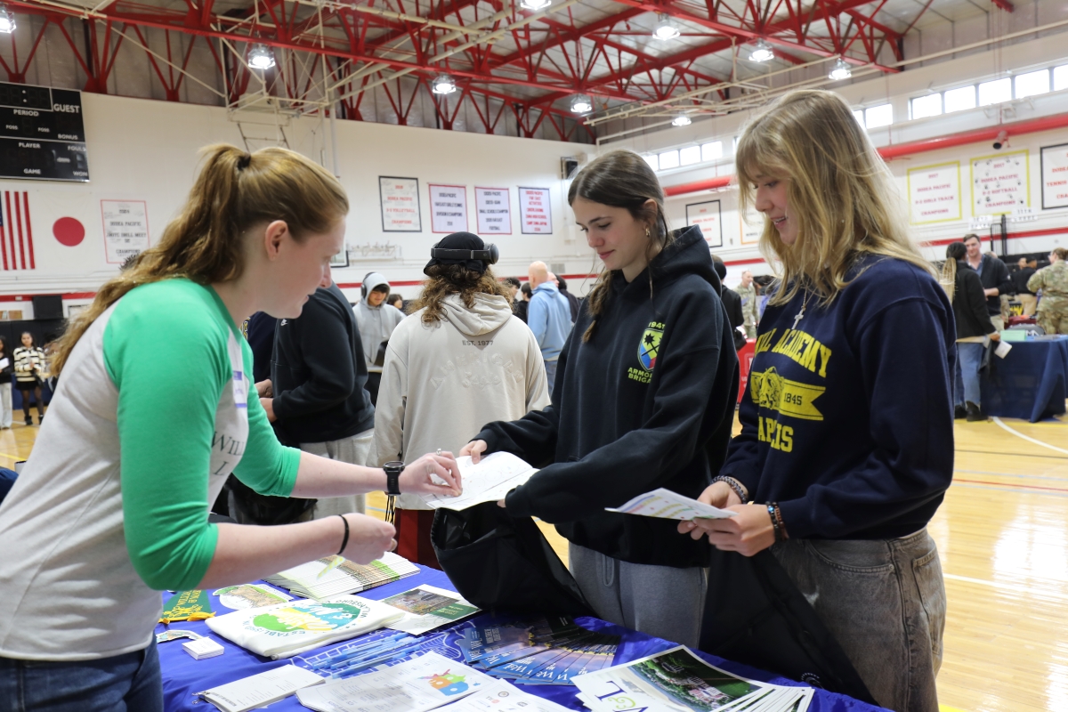 Three teenage girls look over brochures at a school fair booth inside a gymnasium.