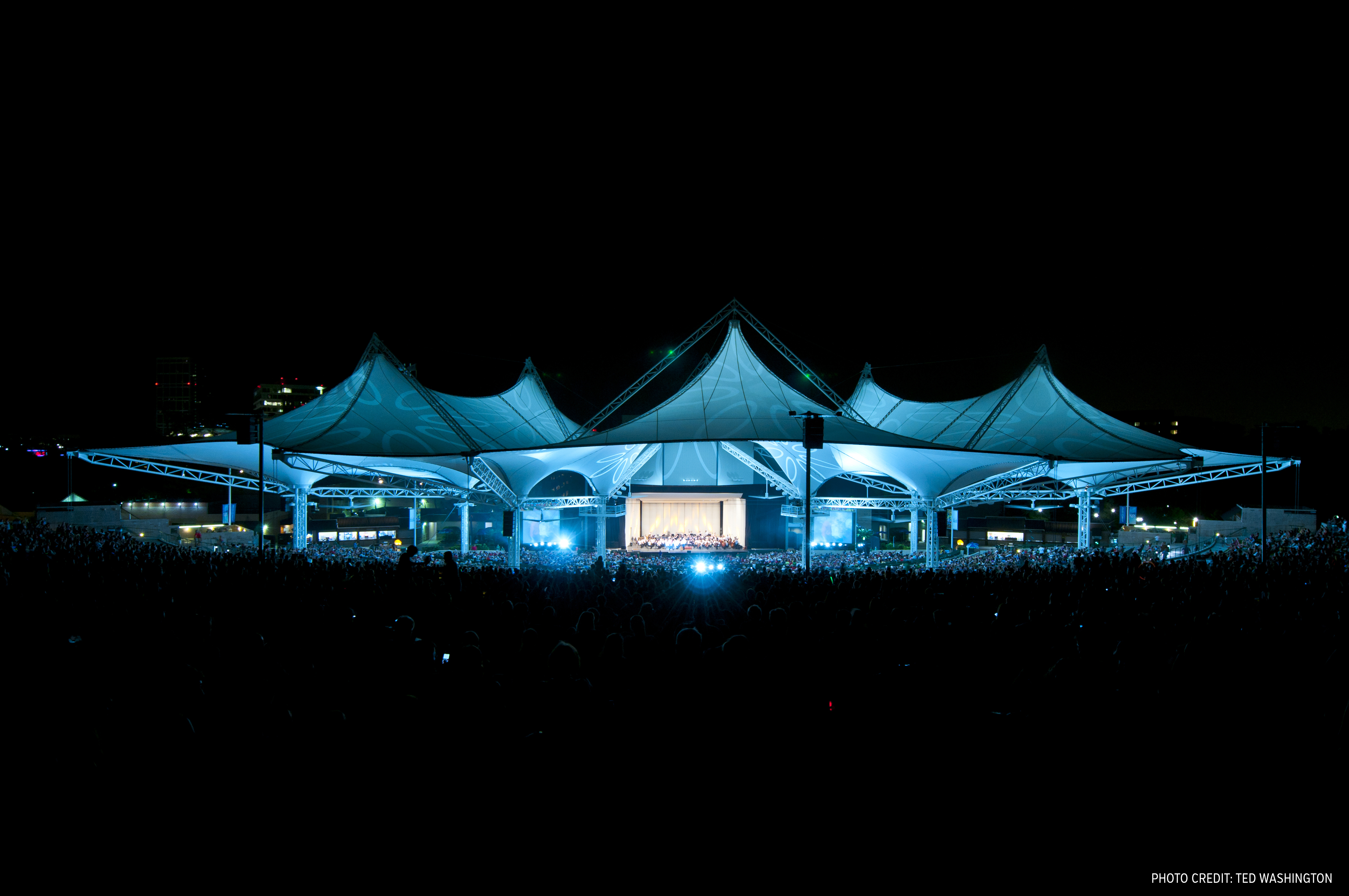 Outdoor amphitheater with tensile roof lit in blue, crowd filling the lawn at night.