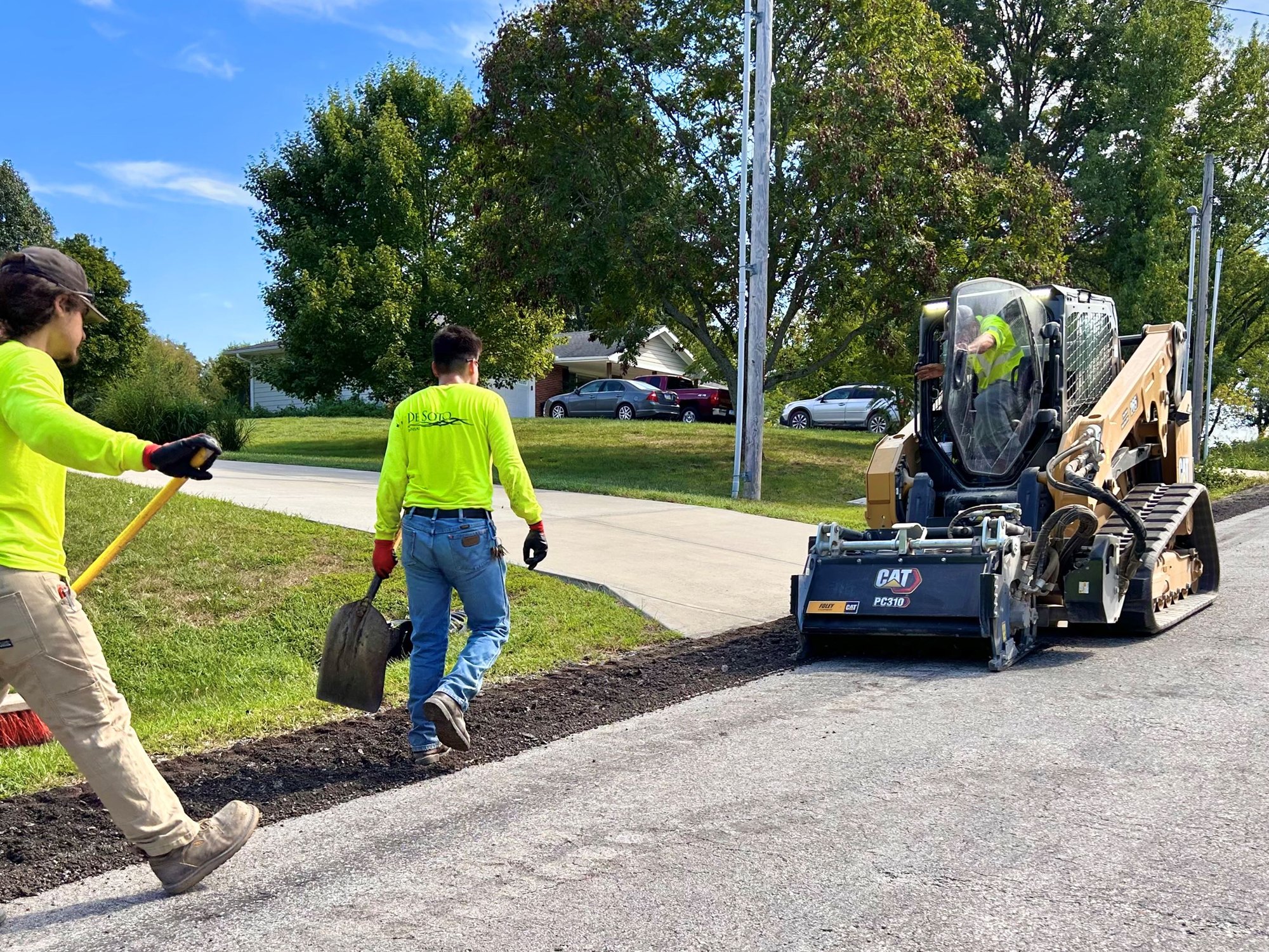 Workers in safety vests and a CAT loader perform asphalt milling on a road surface.
