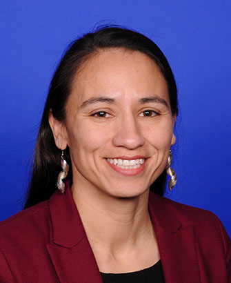 Official portrait of a woman in a dark red blazer smiling against a blue background.
