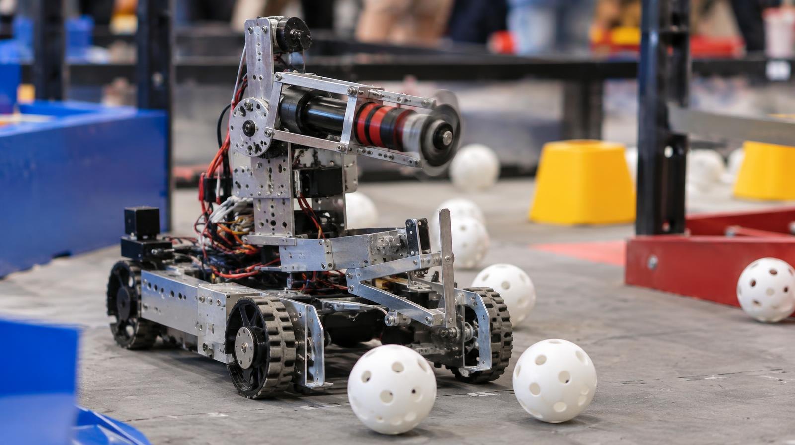 A small competition robot with a turret sits on a game field with white balls nearby.