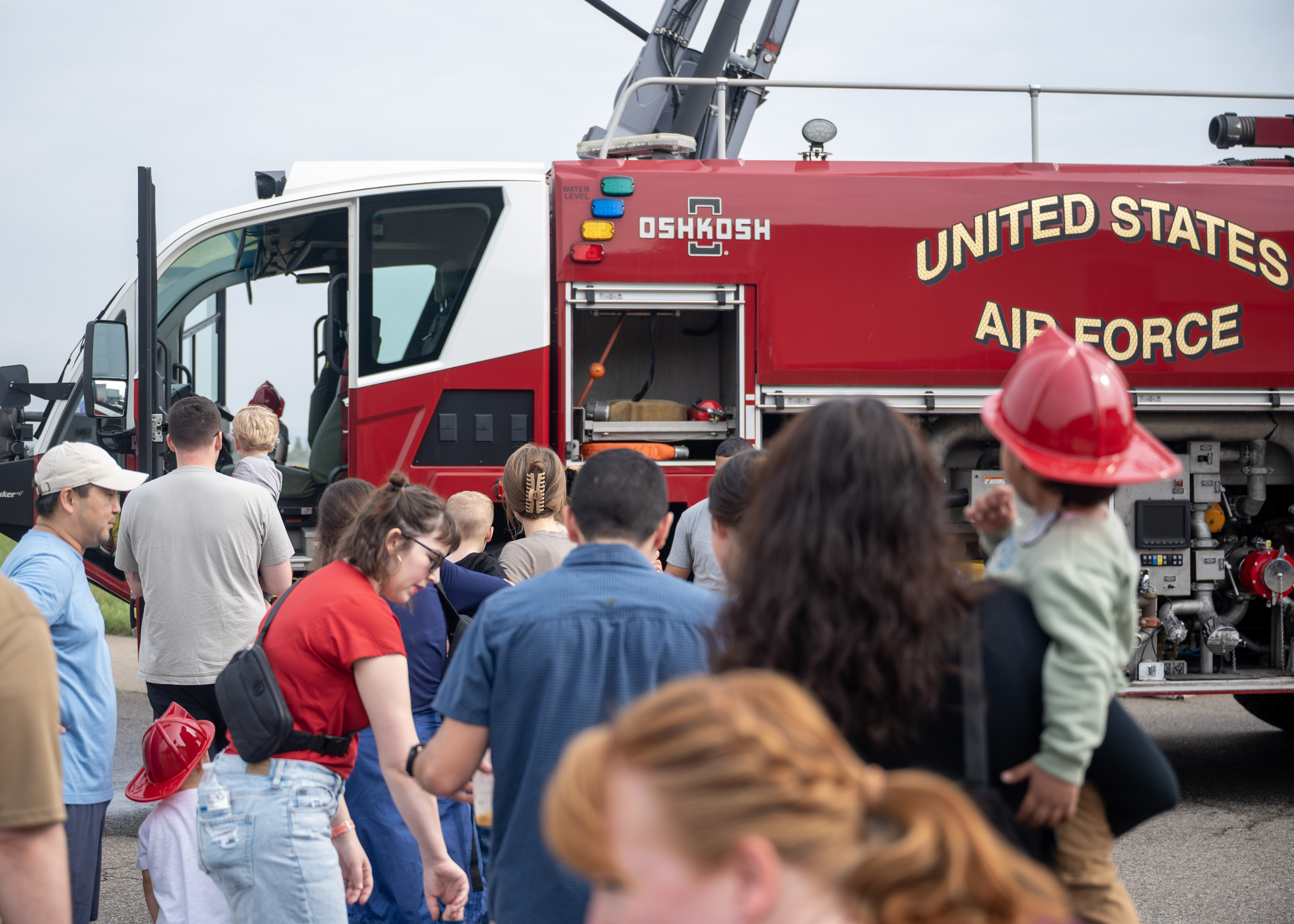 Adults and children line up to view a large red fire truck at an outdoor open house.