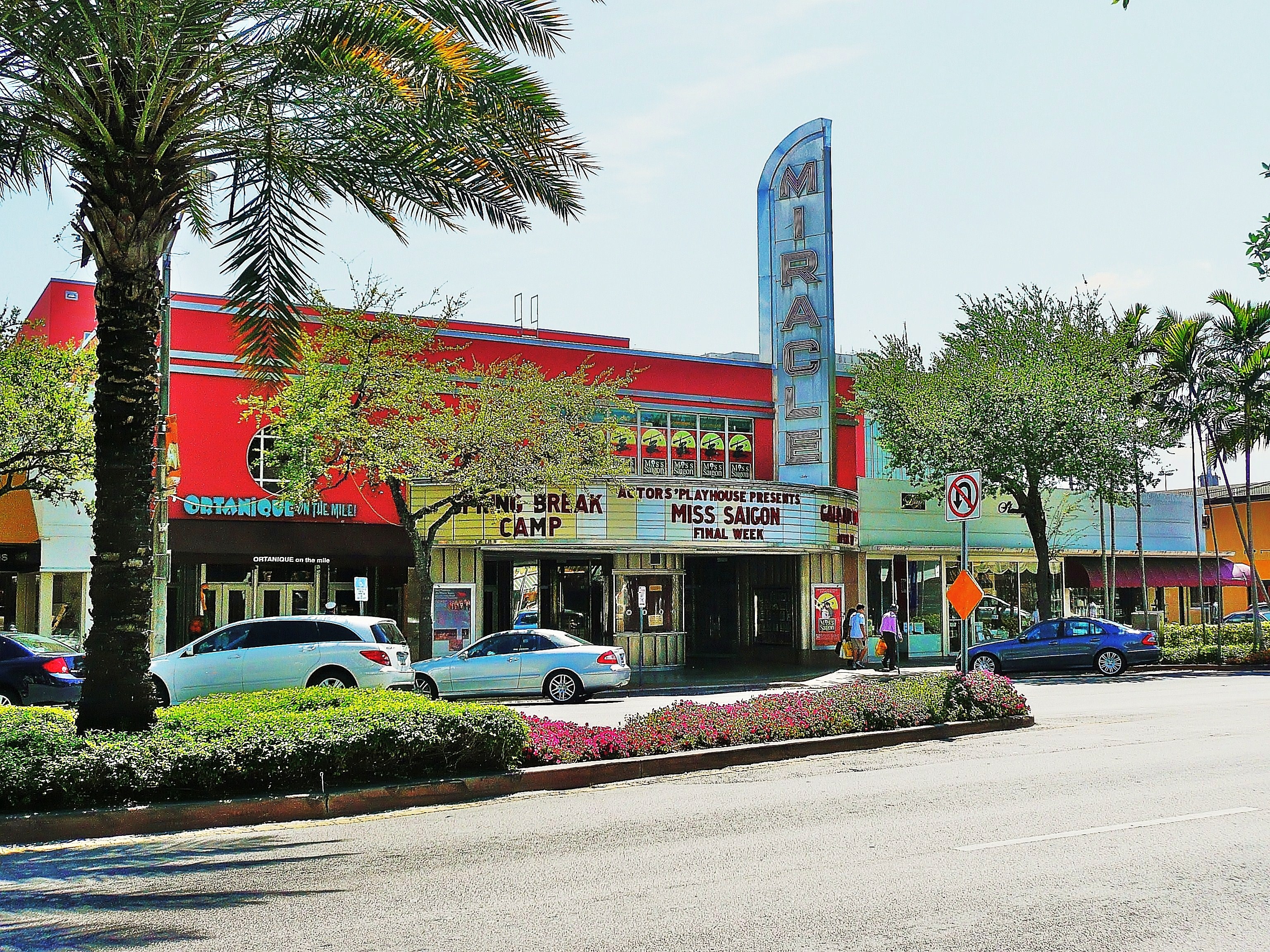 Street-level view of Miracle Mile with a theater marquee, palm trees, and storefronts.