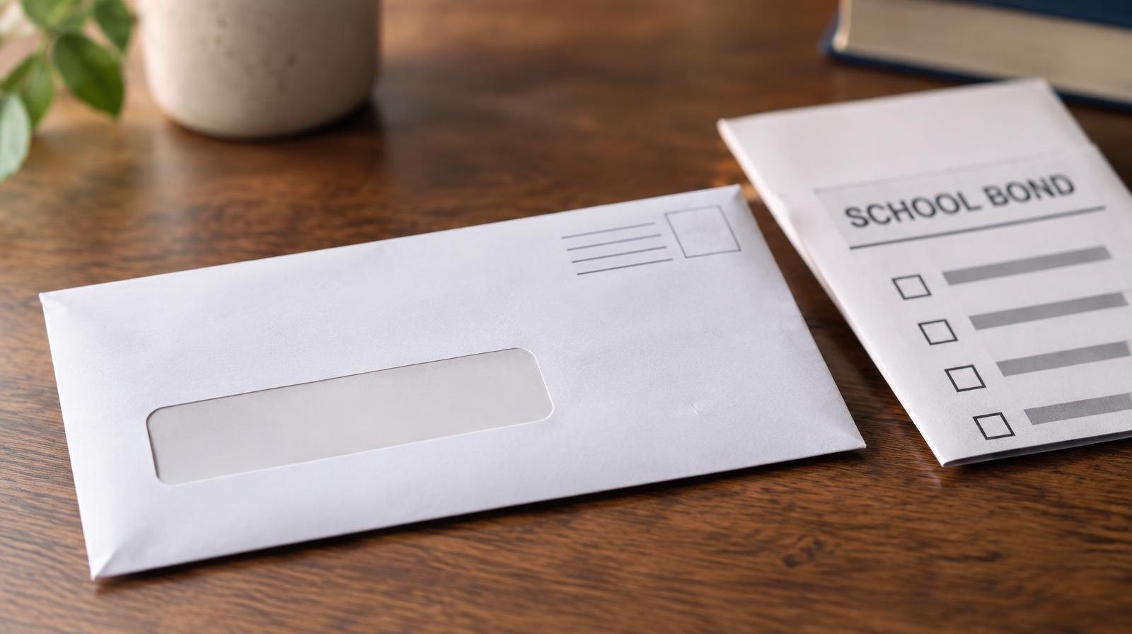 Sealed white vote-by-mail envelope beside a folded paper ballot on a wooden surface.