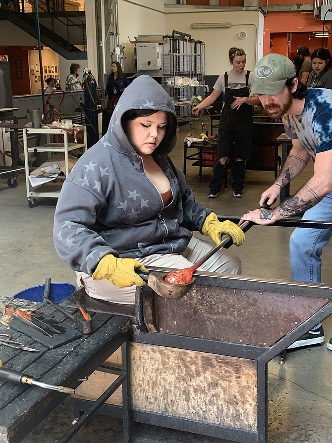 A teen in a hoodie and yellow gloves shapes molten glass at a workbench while an instructor assists.
