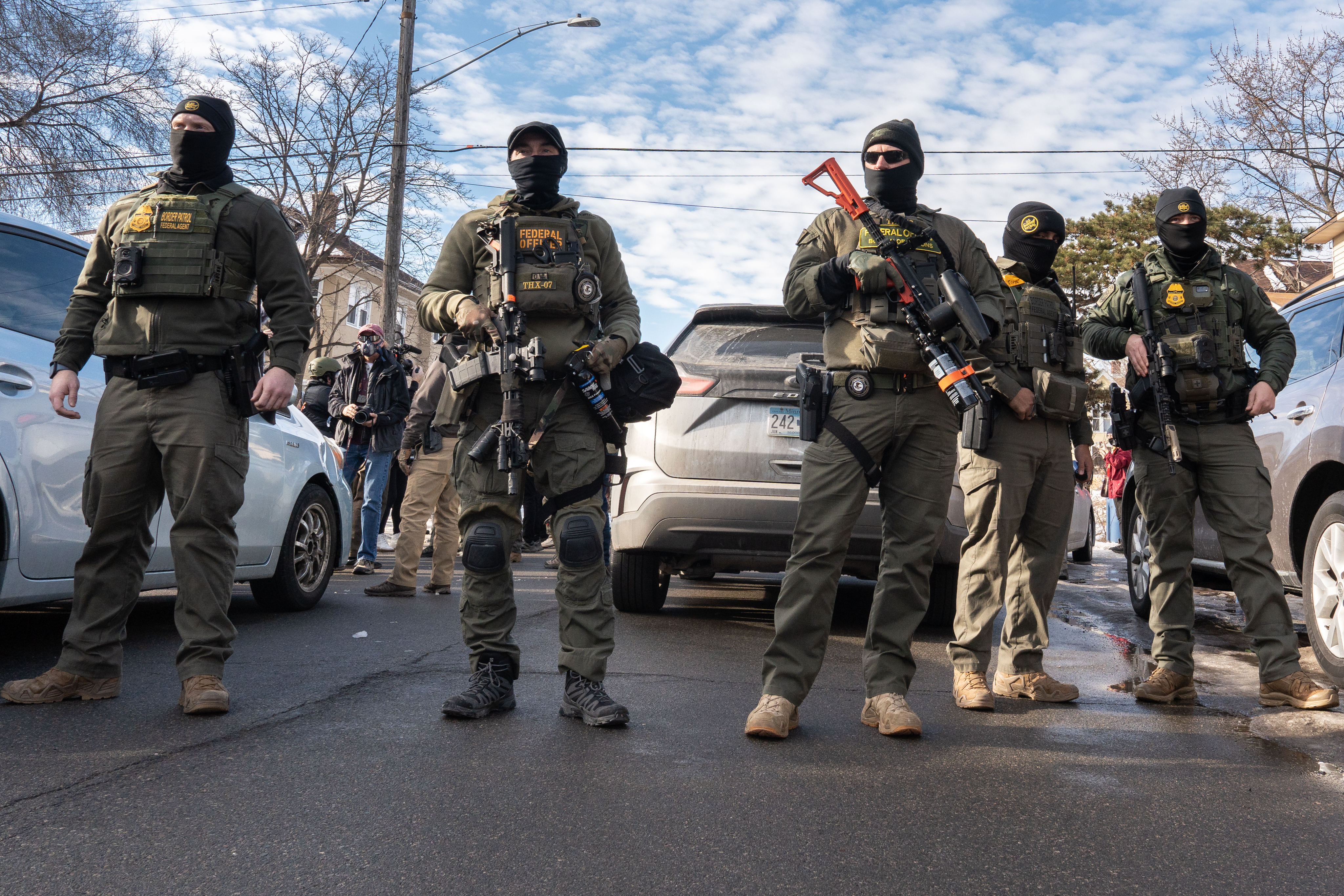 Four armed agents in tactical gear and balaclavas stand on a residential street in winter.
