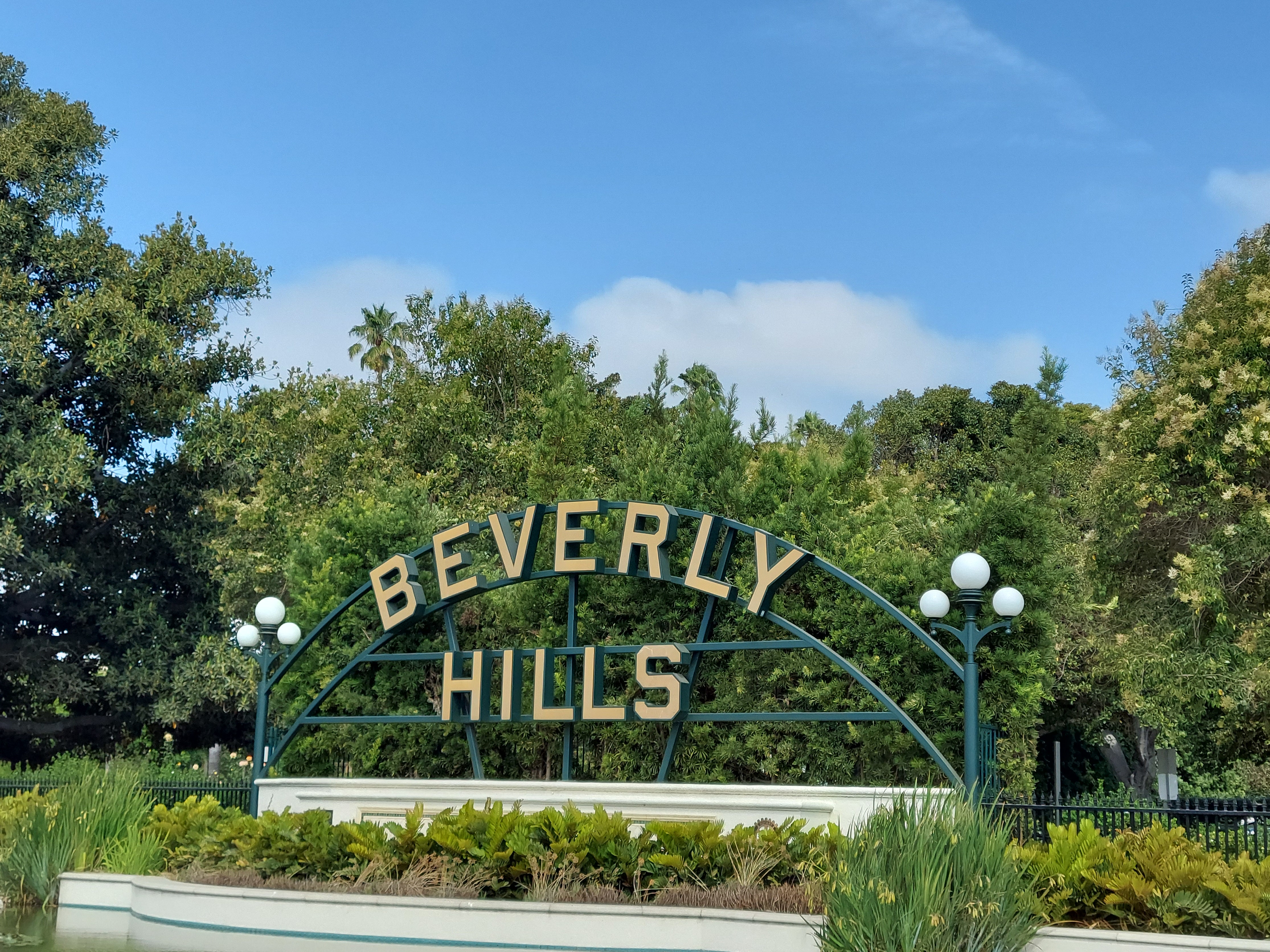 Arched Beverly Hills sign with globe lamp posts surrounded by green trees and lawn.