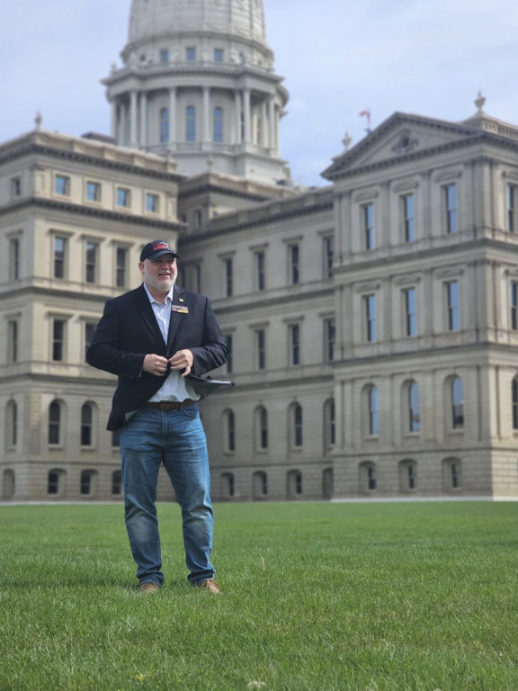 Anthony Hudson stands on a lawn in front of the Michigan Capitol, wearing a dark blazer, jeans and a baseball cap.