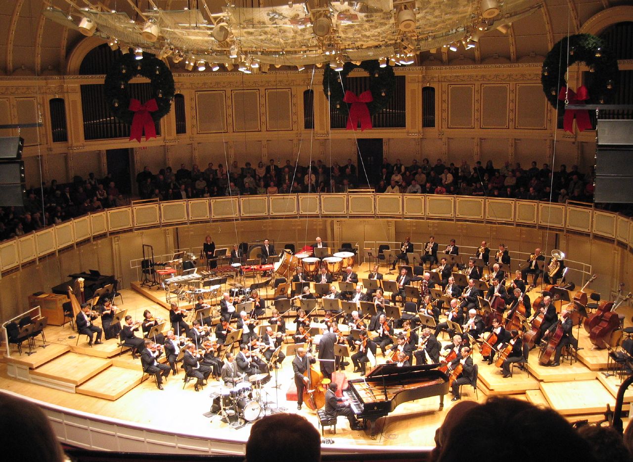 A full symphony orchestra performs on a large concert hall stage with balcony seating above.