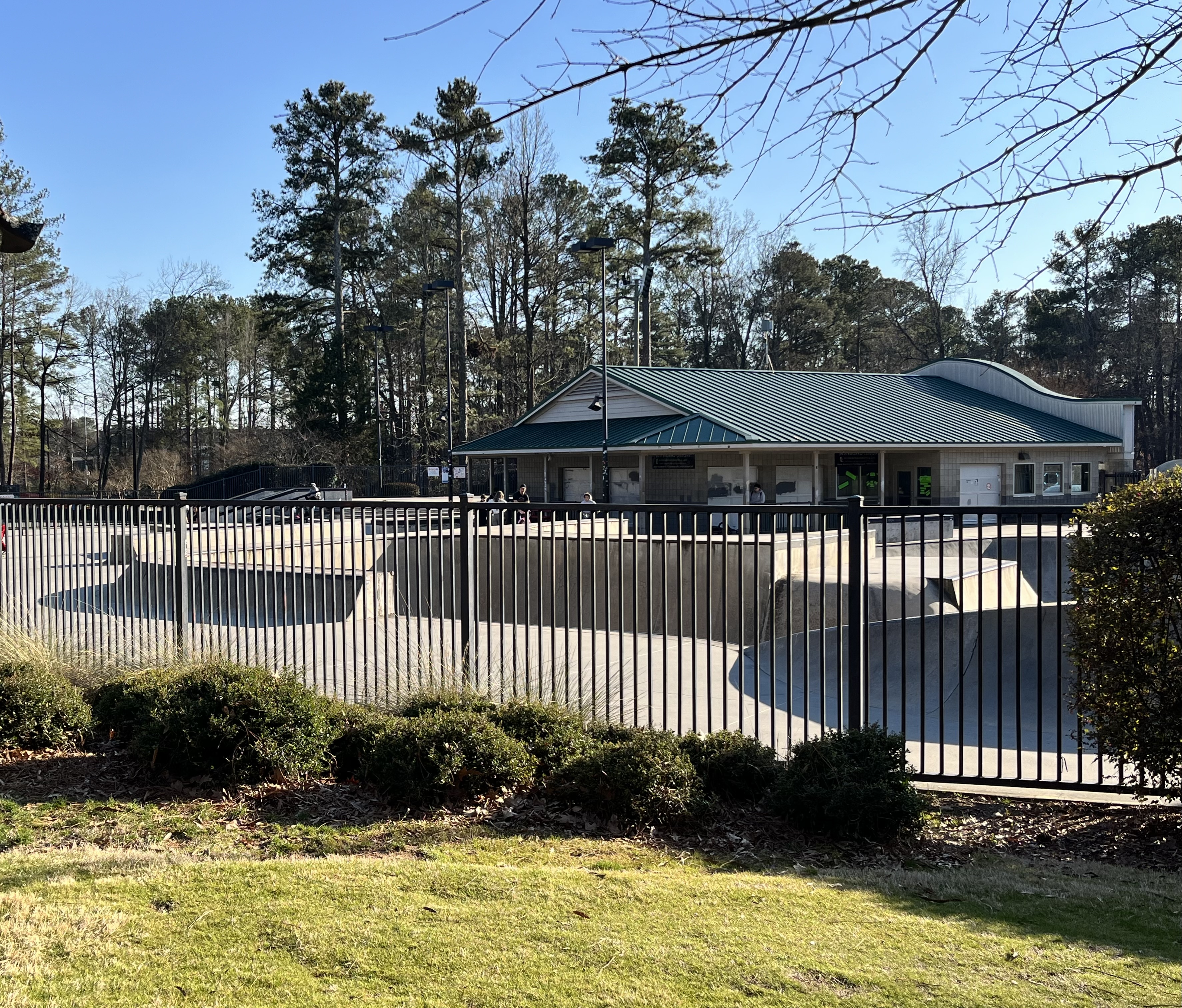 Fenced skate park with a green-roofed building and pine trees on a clear day.