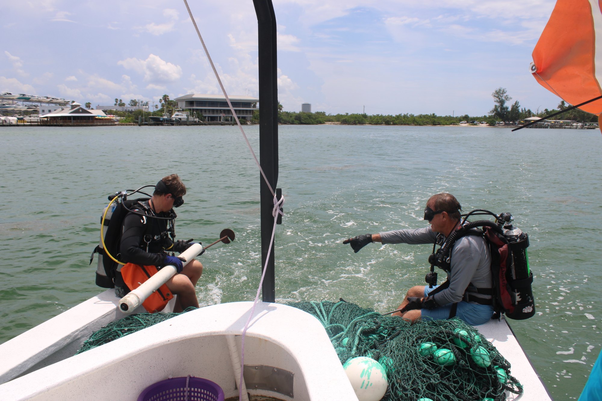Two scuba divers in full gear sit on the stern of a moving boat on a calm bay.