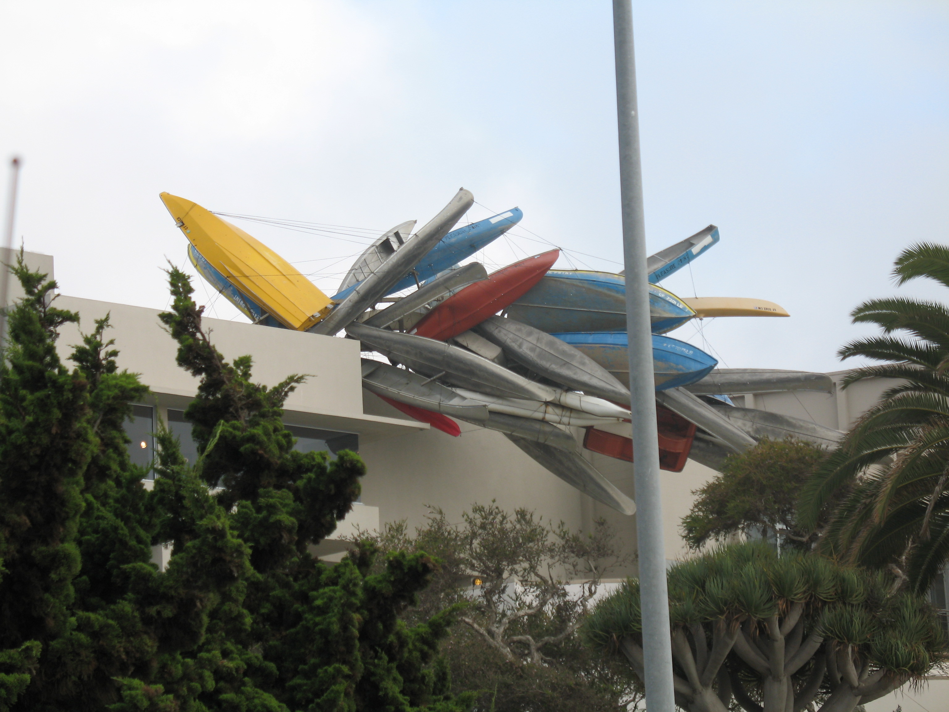 Large colorful sculpture of kayaks and watercraft projecting from a museum rooftop, surrounded by trees.